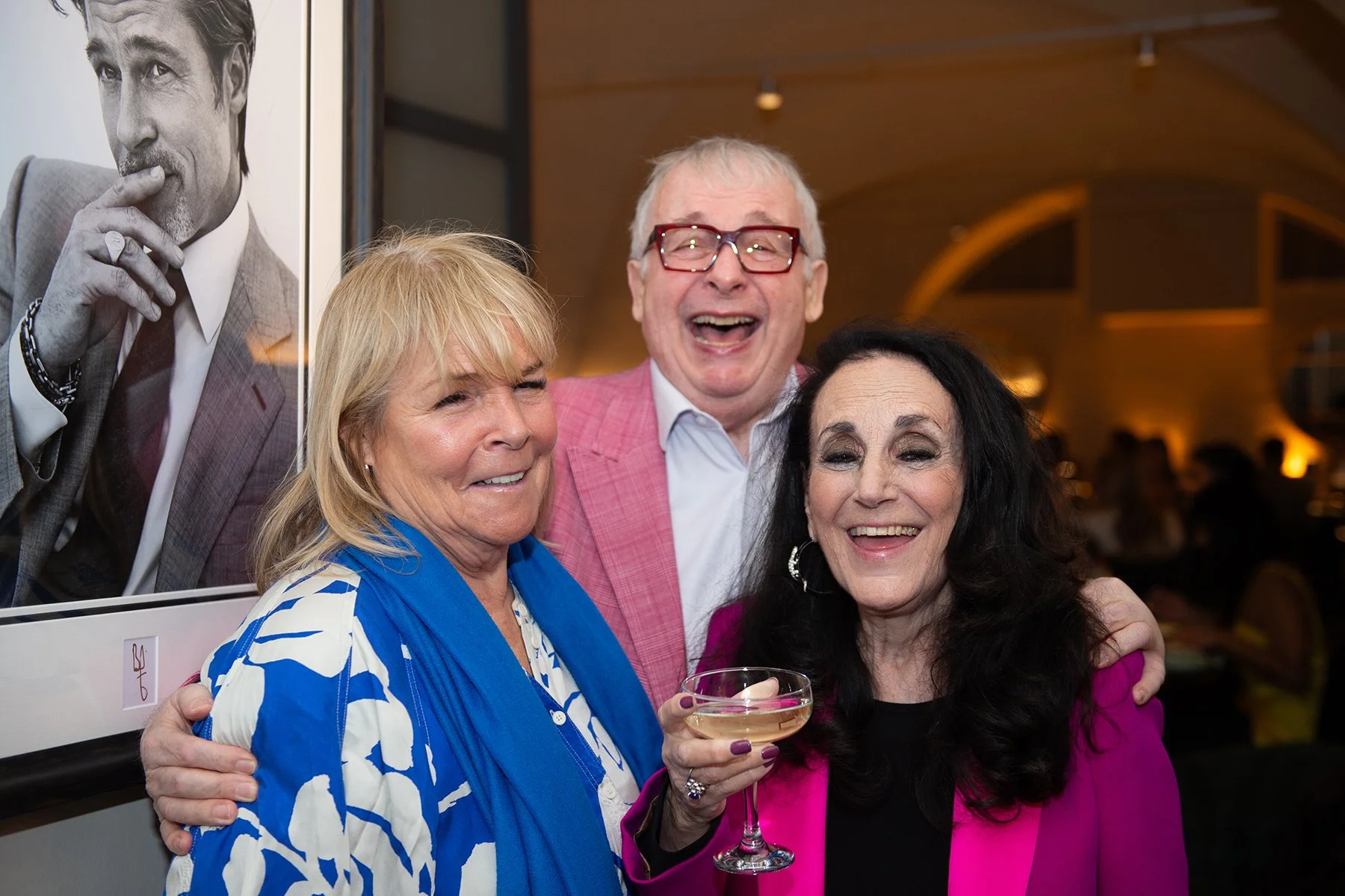 Three people smiling and posing together at a social event, one woman holding a glass of white wine, with a black-and-white portrait of a man in the background.