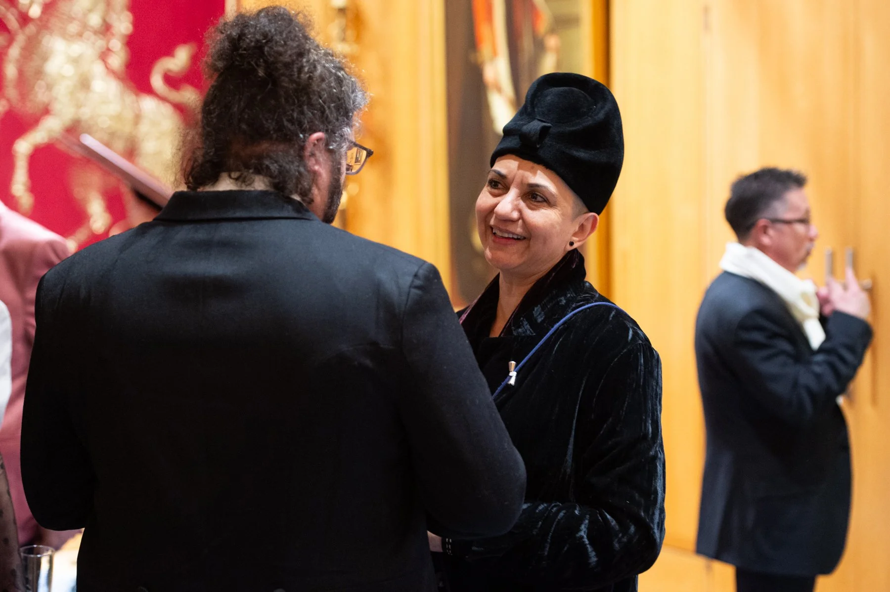 Two people are talking and smiling at each other at an event. The woman is wearing a black hat and velvet blazer, and the man has curly hair and glasses. There are gold and red decorations in the background, with other individuals in the background, 