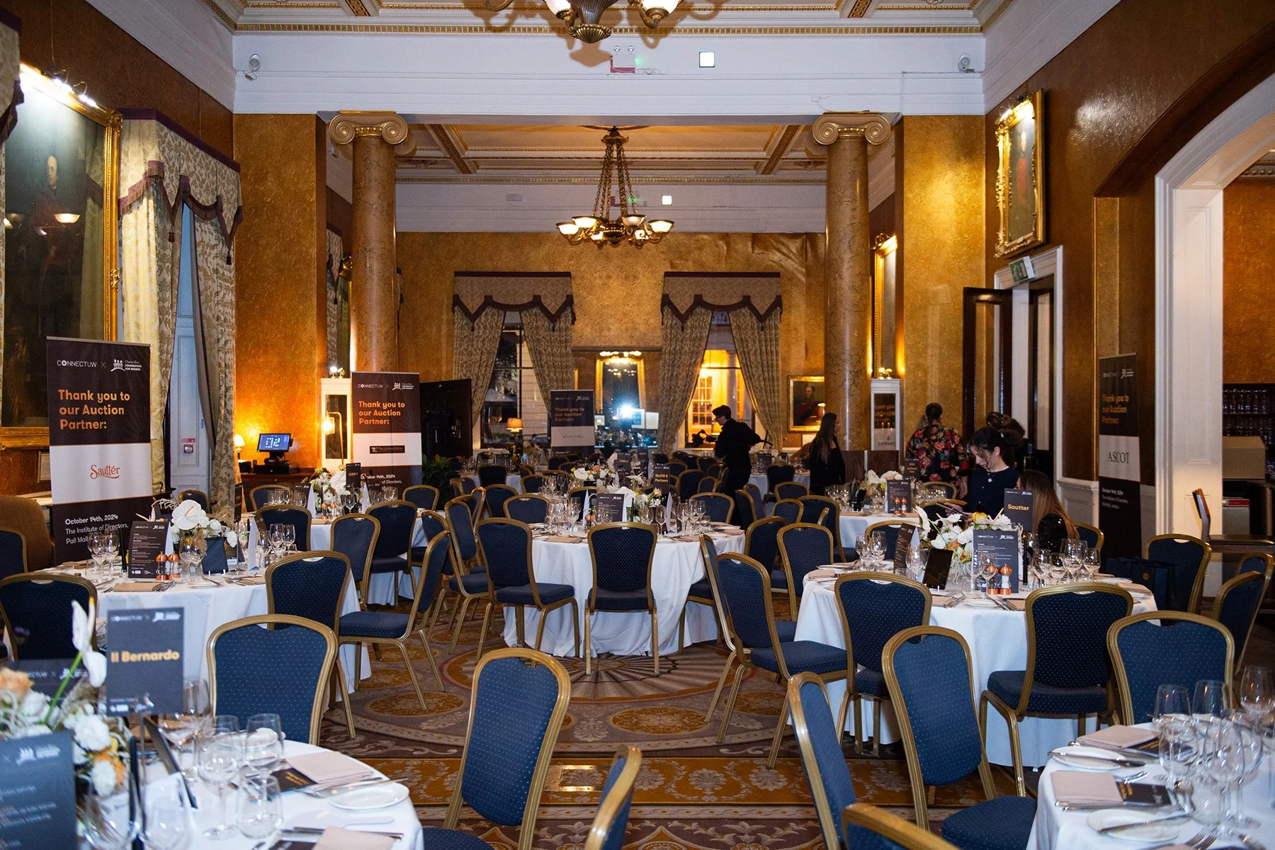 A large banquet room decorated for an event with multiple round tables covered in white tablecloths and set with glassware, silverware, and floral centerpieces. The room features tall windows with elaborate curtains, a high ceiling with chandeliers, 