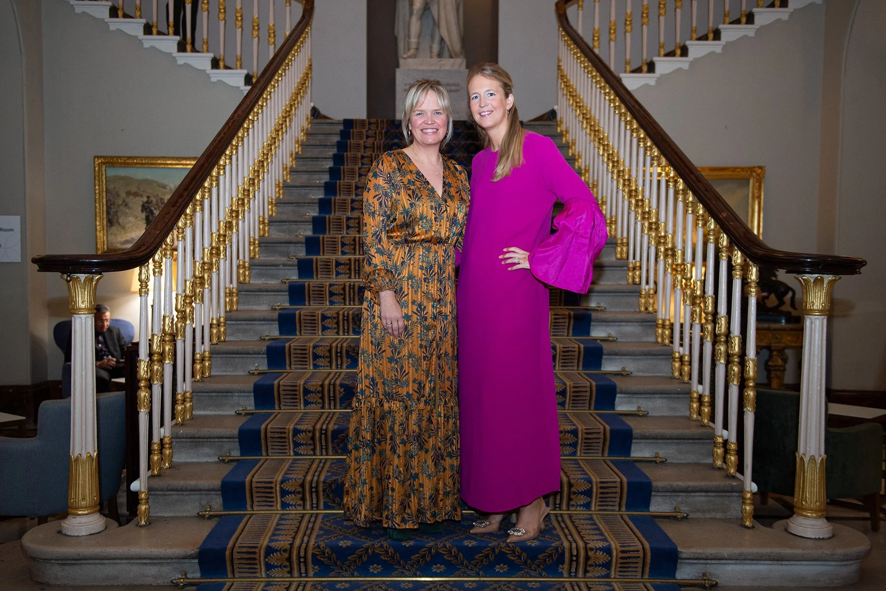 Two women standing on a grand staircase with ornate white and gold railings. One woman is wearing a floral, long-sleeved dress, and the other is in a bright pink gown with bell sleeves. They are smiling and posing for the photograph inside a formal b