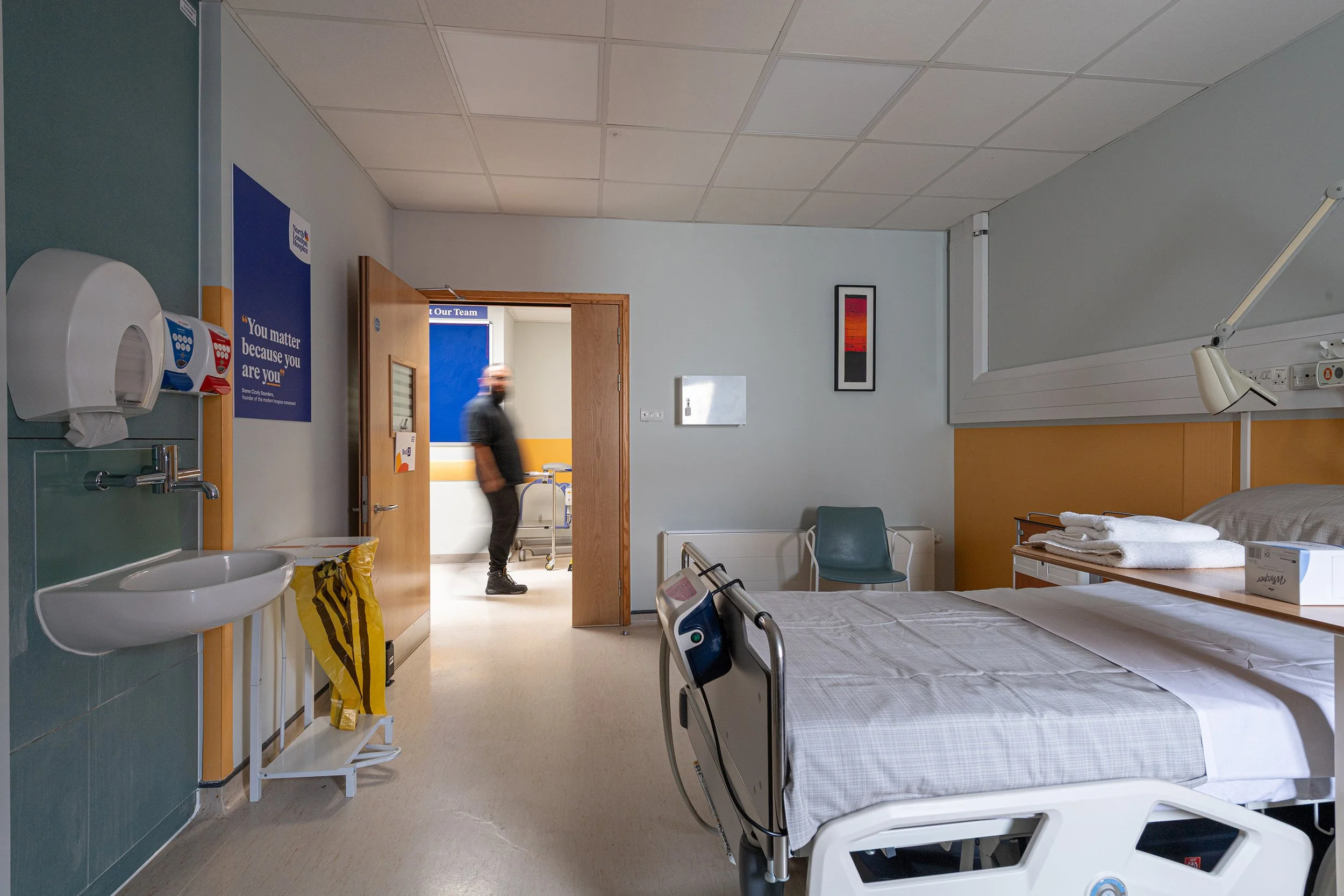 A hospital room with two empty hospital beds, a chair, a sink with soap dispenser, a yellow bag, and a person standing in the doorway. The room has white walls with some pictures and medical equipment.