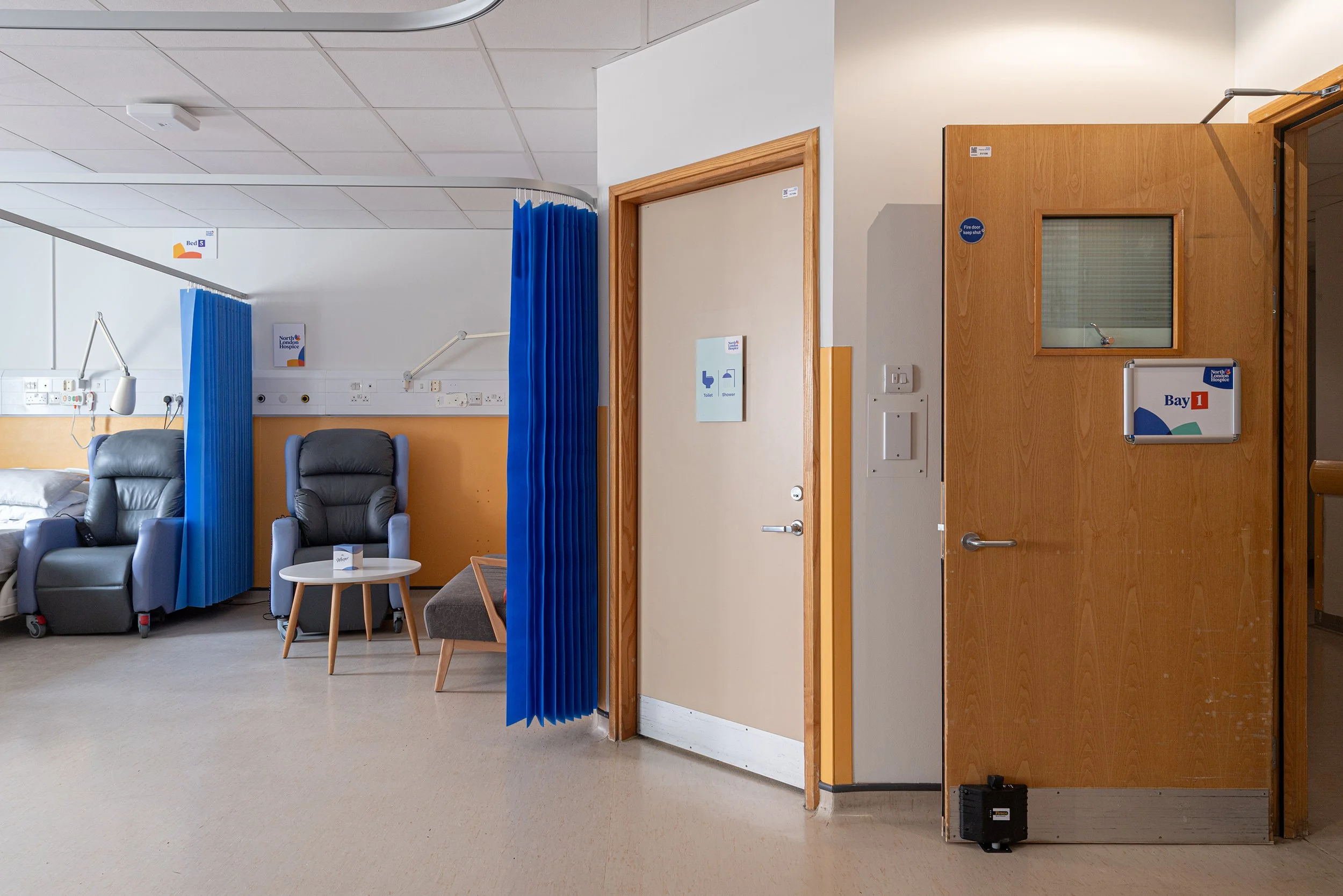 Hospital room with two reclining chairs, a small table, medical equipment, and a privacy curtain; doorway with signage indicating bathroom facilities.