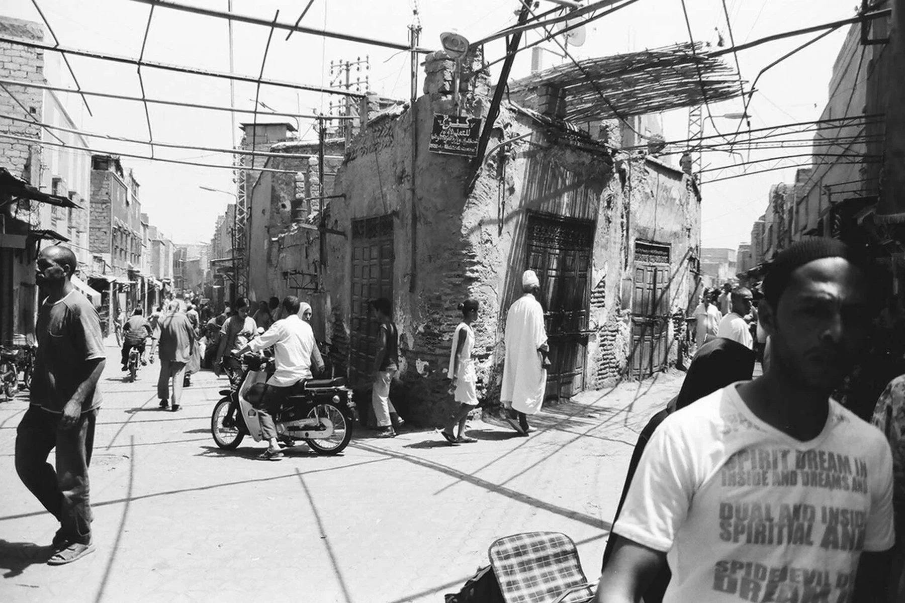A busy street scene in an urban area with people walking and riding bicycles. Old, damaged buildings with exposed brick and scaffolding are visible, and electrical wires are overhead. The image is in black and white.