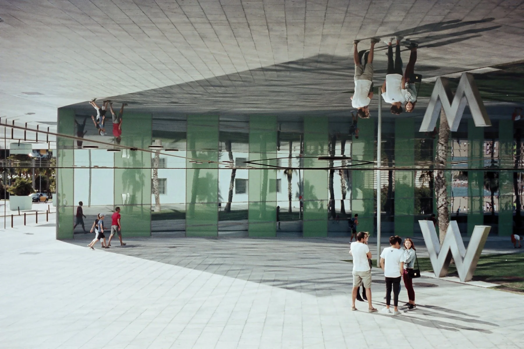 People walking and talking in front of a modern building with reflective glass walls and large metal letters. The image is upside down.