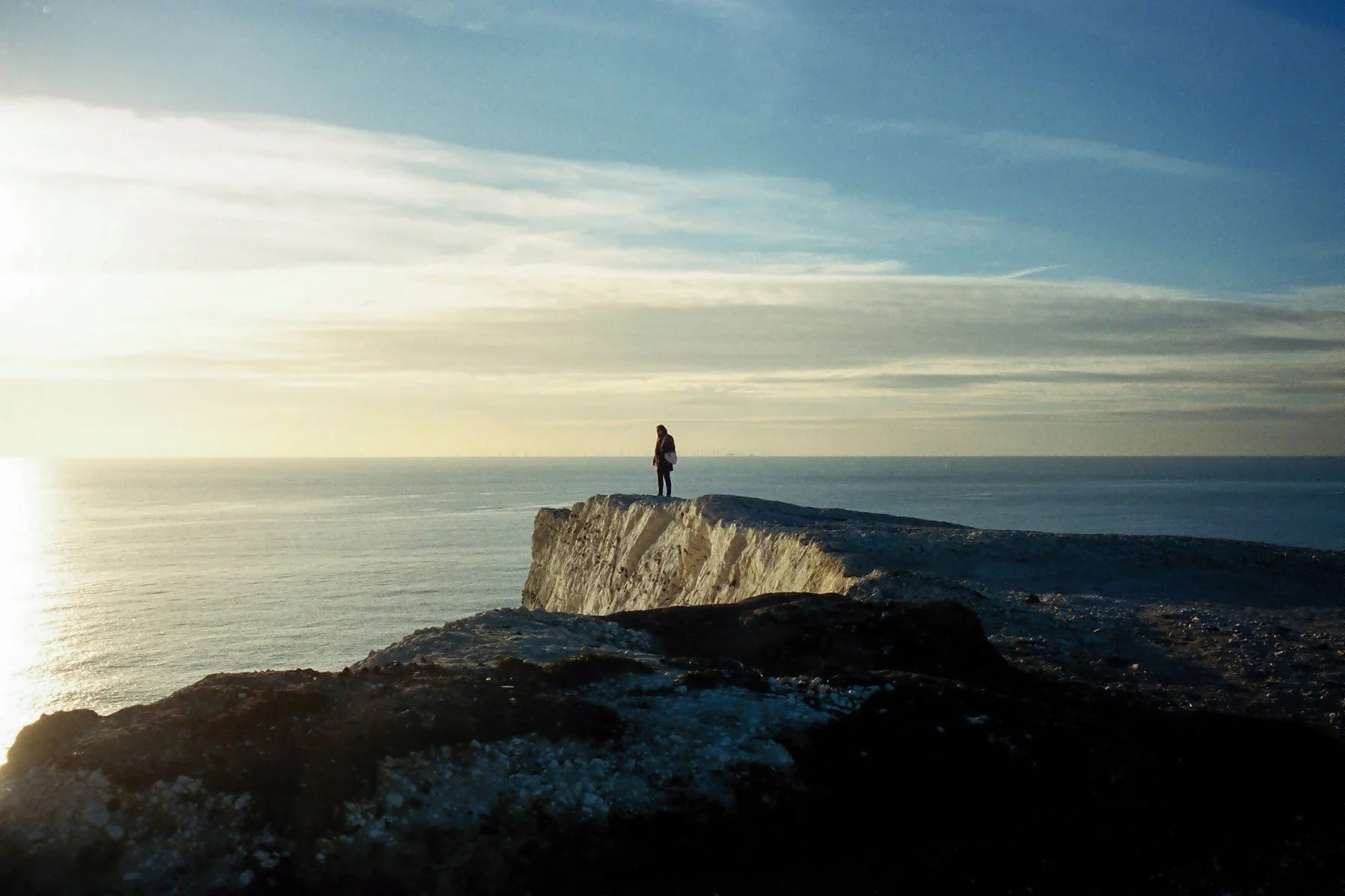 A person standing on the edge of a rocky cliff overlooking the ocean during sunset.