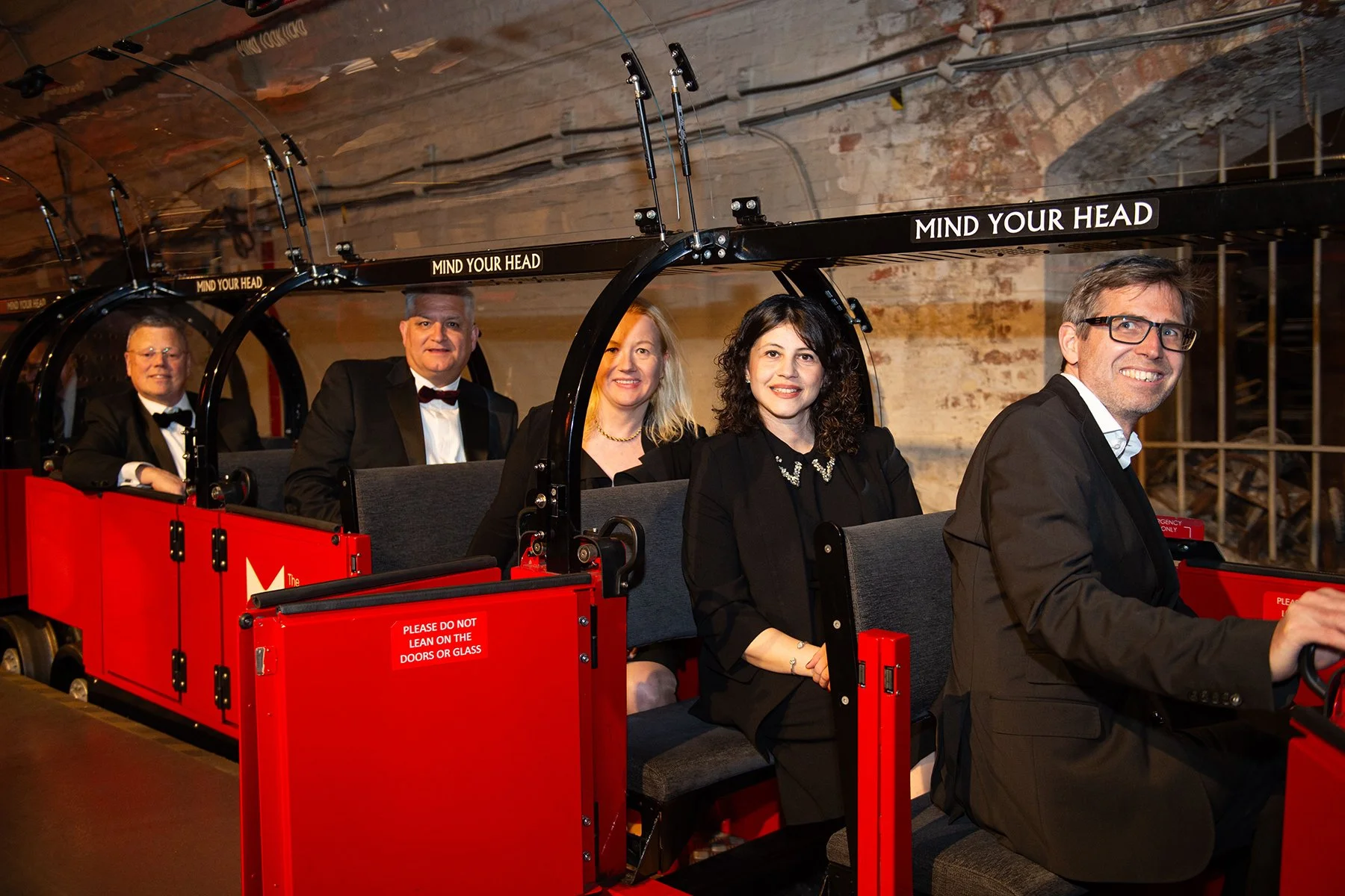 People riding a red tram or trolley with black frames in an indoor setting, with brick walls and wooden ceiling beams in the background, all dressed in formal attire.