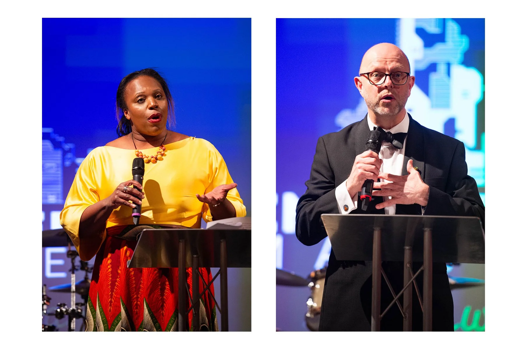 Two speakers, a woman in a yellow dress with red and green skirt and beads, and a man in a black tuxedo with a bow tie, speaking into microphones at podiums during an event.