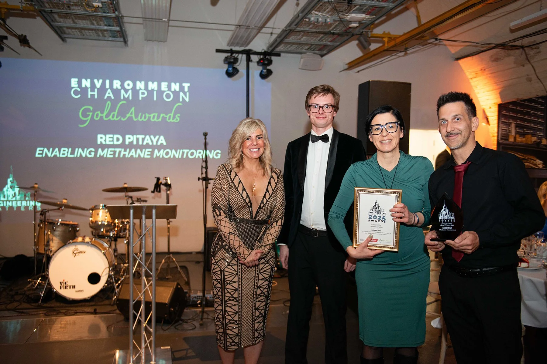 People at an award ceremony with a stage, musical instruments, and a large screen displaying awards for environment champion, Gold Awards, Red Pitaya, and enabling methane monitoring. Four individuals are in the foreground, three women and one man, h