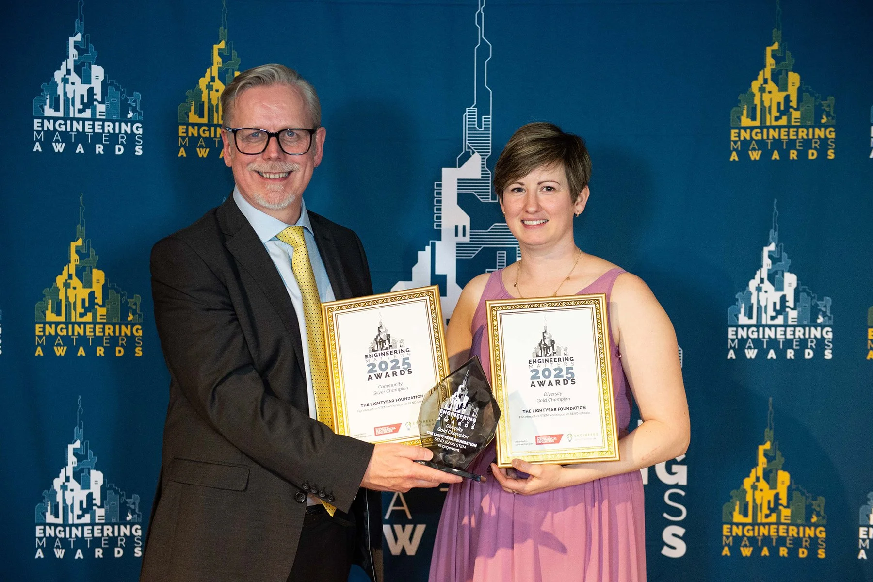 Two individuals smiling and holding awards at the Engineering Matters Awards event. The man on the left is wearing glasses, a black suit, light blue shirt, and yellow tie. The woman on the right is wearing a purple dress. Both are holding framed cert