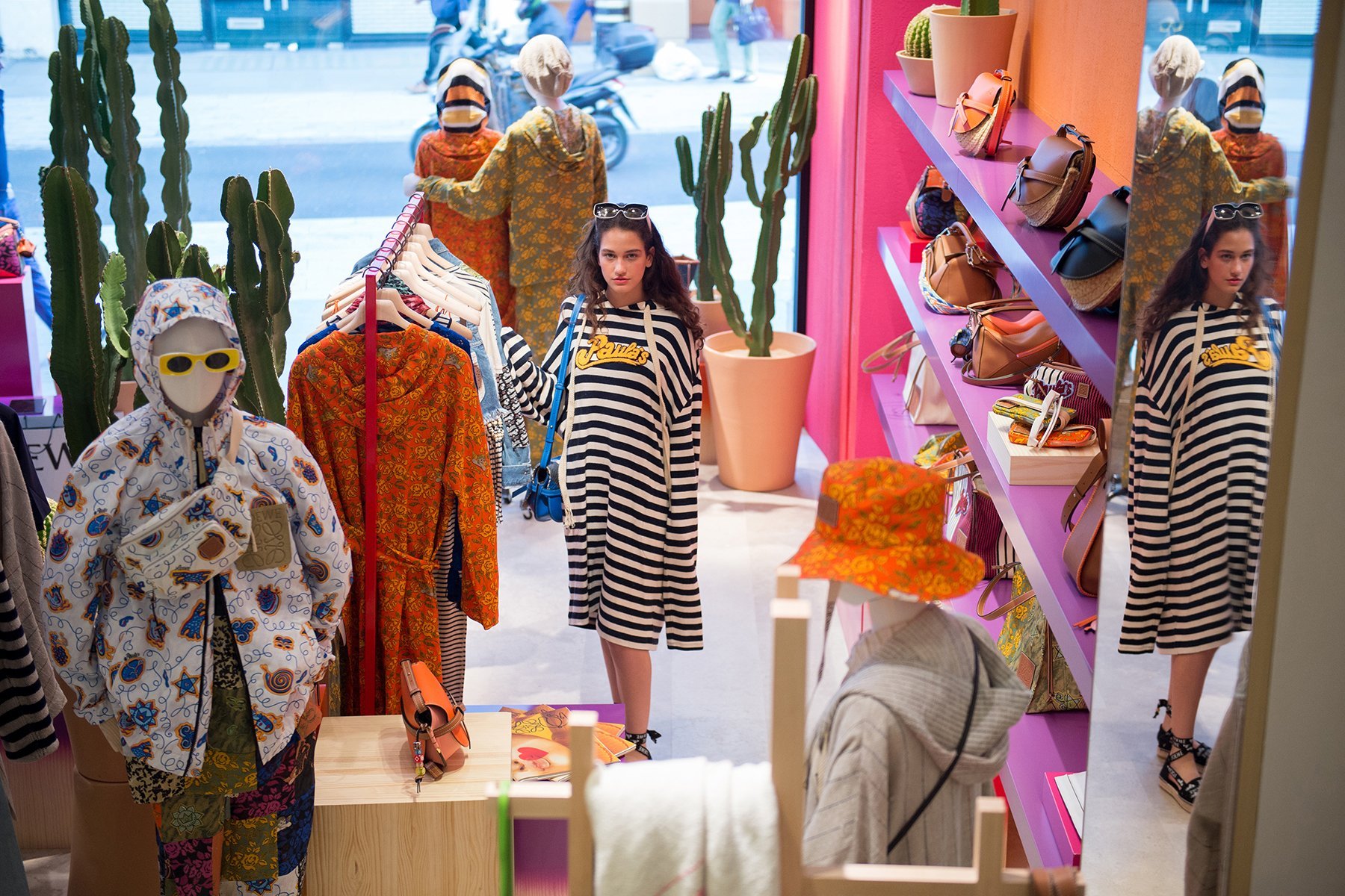 Interior of a clothing store with mannequins wearing colorful clothing, including a woman in a black and white striped dress, and shelves displaying handbags and accessories, with large cactus plants near the window.