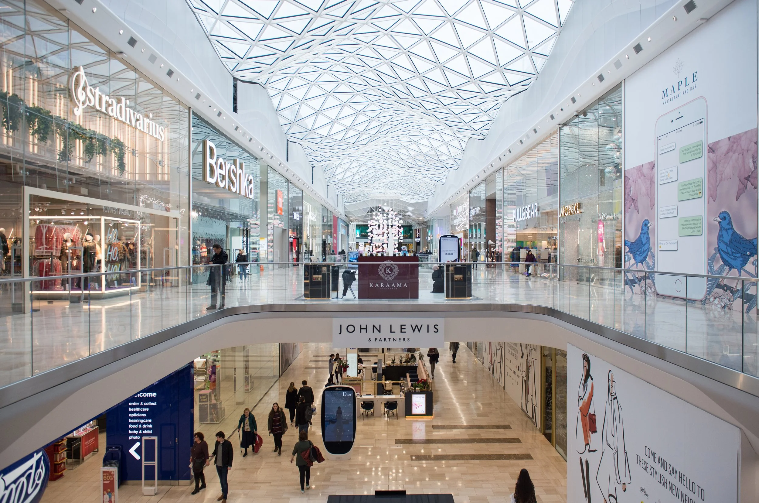 Interior of a modern shopping mall with multiple levels, crystal-like ceiling design, various store signs such as Estradivarius, Bershka, and others, with shoppers walking and browsing.