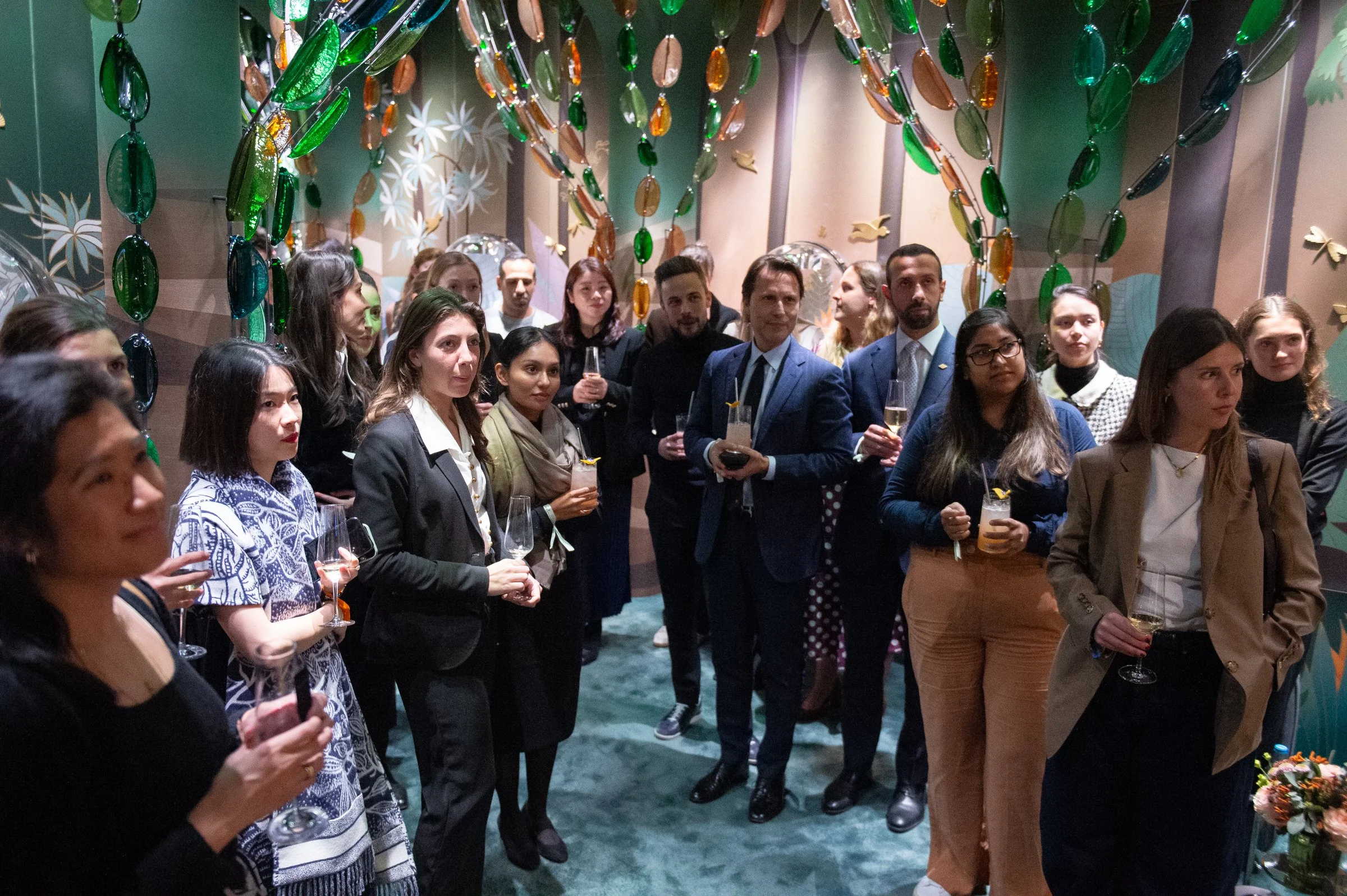 A group of people dressed in business or semi-formal attire gathered at an indoor event, holding glasses of drinks. The setting features decorative elements like hanging glass ornaments and artwork on the walls, indicating a festive or celebratory at