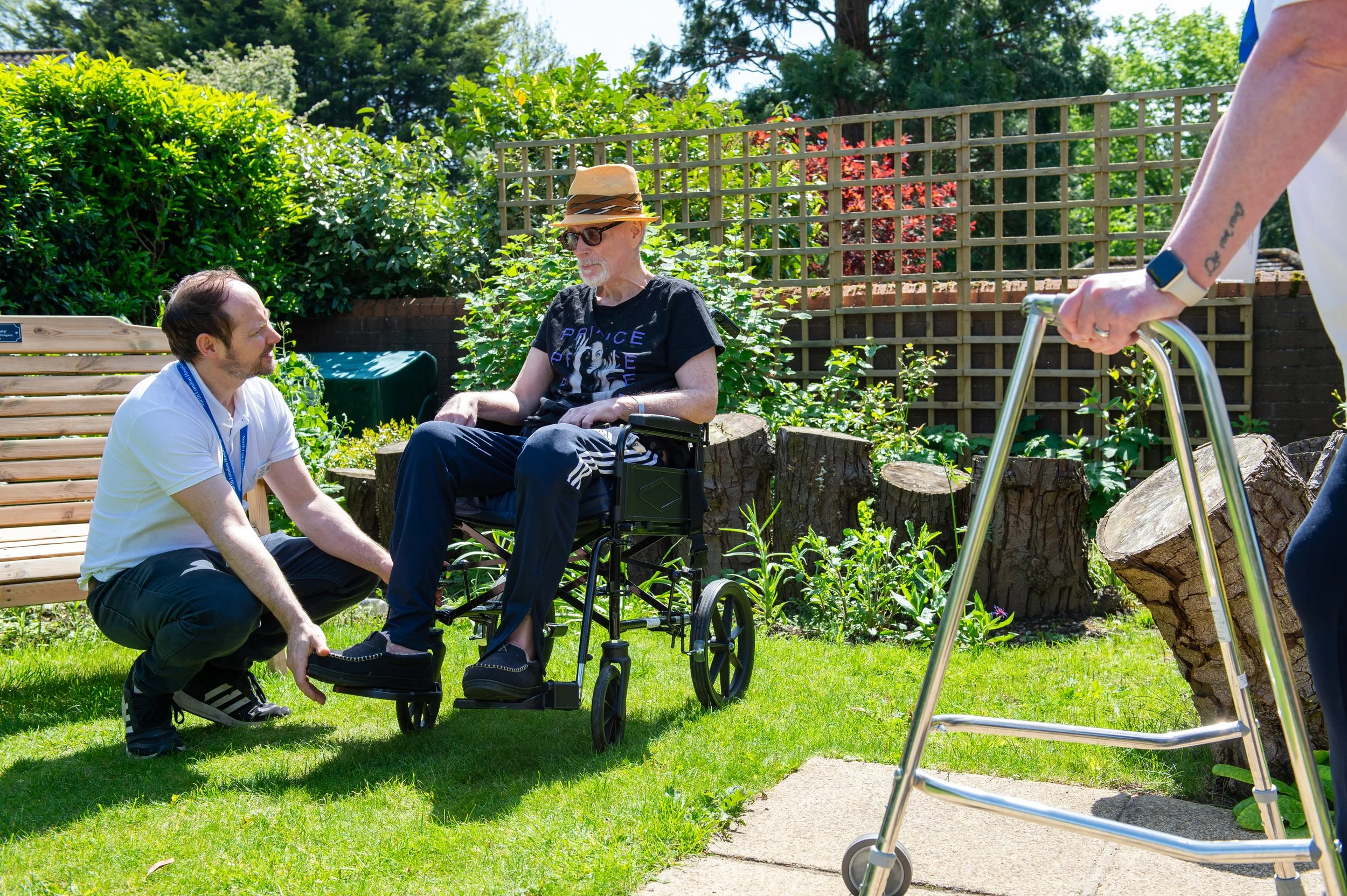 A man kneeling on the grass talking to an elderly man in a wheelchair outdoors on a sunny day, with a woman holding a walker in the foreground.