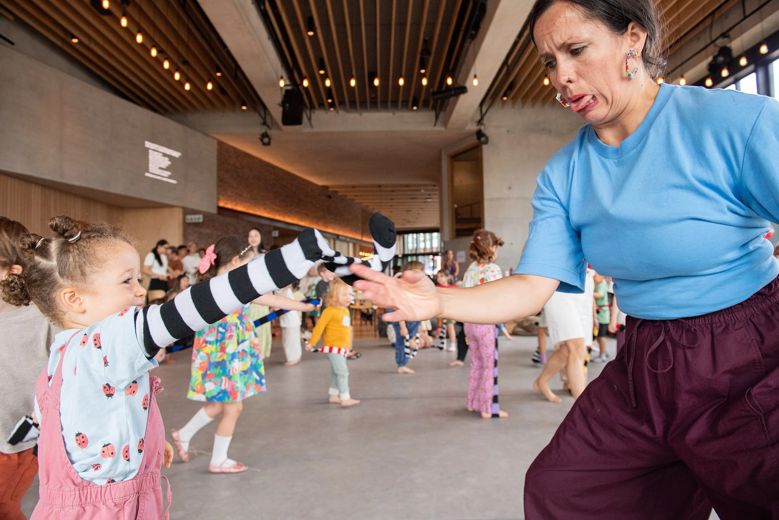 Young girl reaching out to a woman during a dance activity in a large indoor space filled with children.
