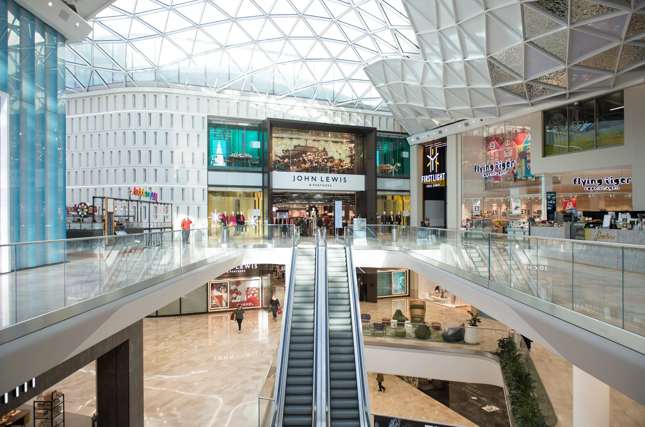The interior of a modern shopping mall with multiple levels, escalators, and storefronts including 'John Lewis & Partners' and 'Flying Tiger Copenhagen', featuring a glass and steel dome ceiling letting in natural light.