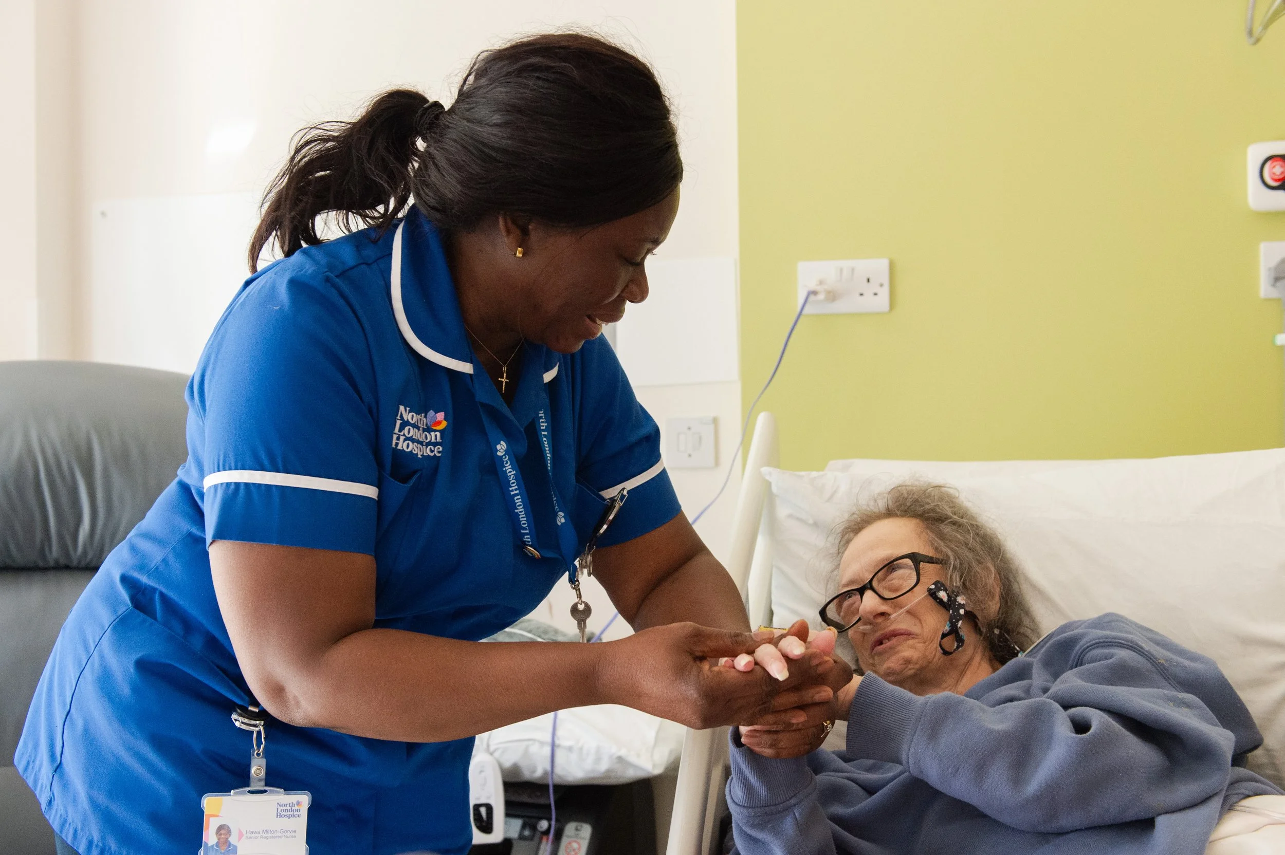 A healthcare worker, dressed in blue uniform with North London Hospice badge, is caring for an elderly patient lying in bed. The nurse is holding the patient's hand, showing compassion and support in a hospital or hospice room.