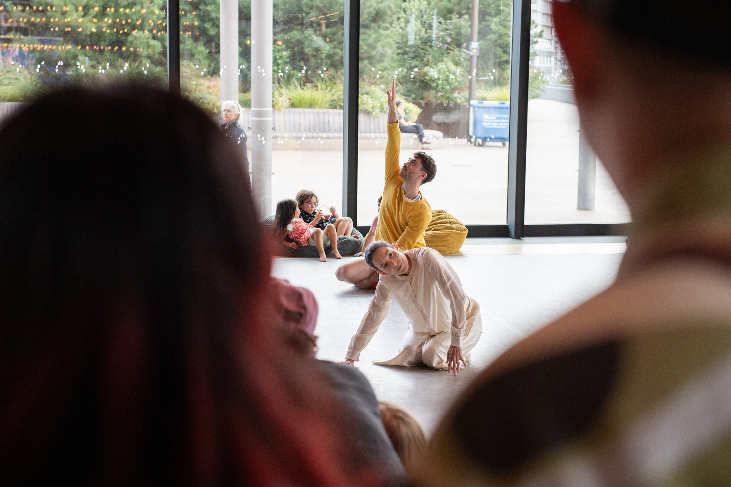 People, including children and adults, practicing yoga or stretching exercises inside a modern room with large glass windows, outdoor trees, and string lights visible outside.