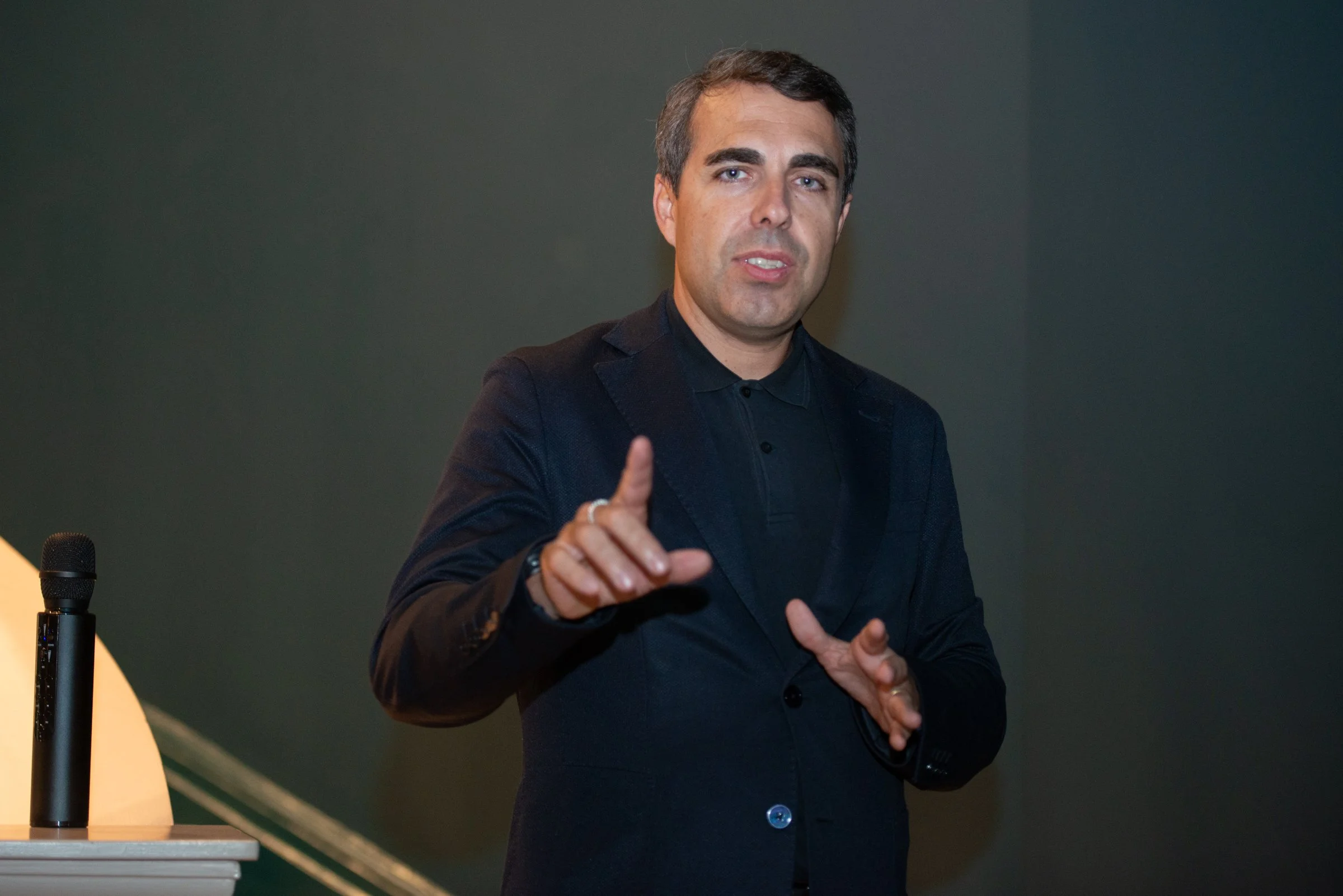 A man in a black suit and black shirt speaking at what appears to be a conference or seminar, gesturing with his right hand.