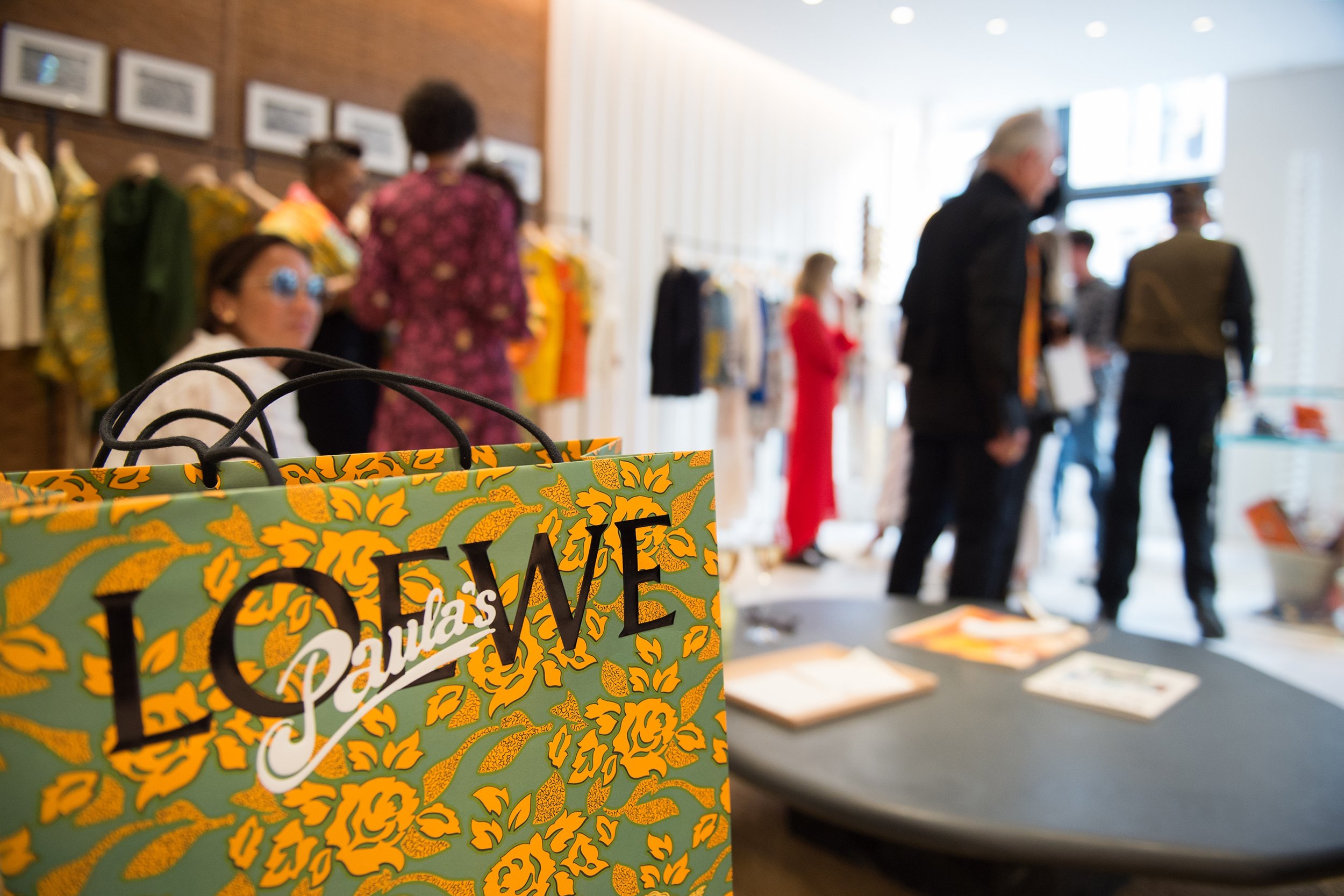Bright yellow and green shopping bag with floral design, partially visible, with black handles, in front of a blurred retail store or exhibit with people and clothing displays.