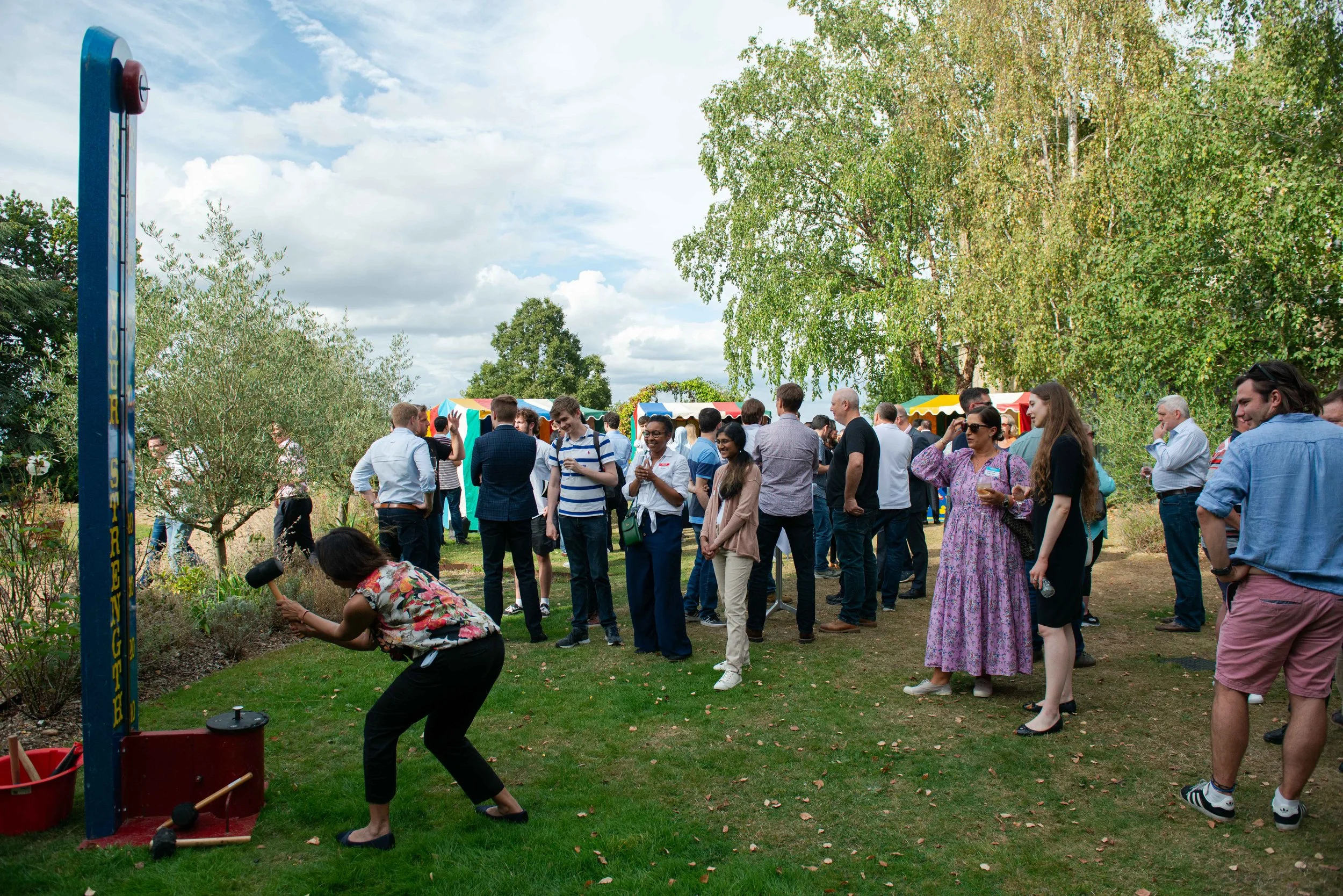 People gathered outdoors at a festival or fair, with colorful tents and large trees in the background. A woman in a floral top is playing a game involving hitting objects with a hammer, while others watch and converse.