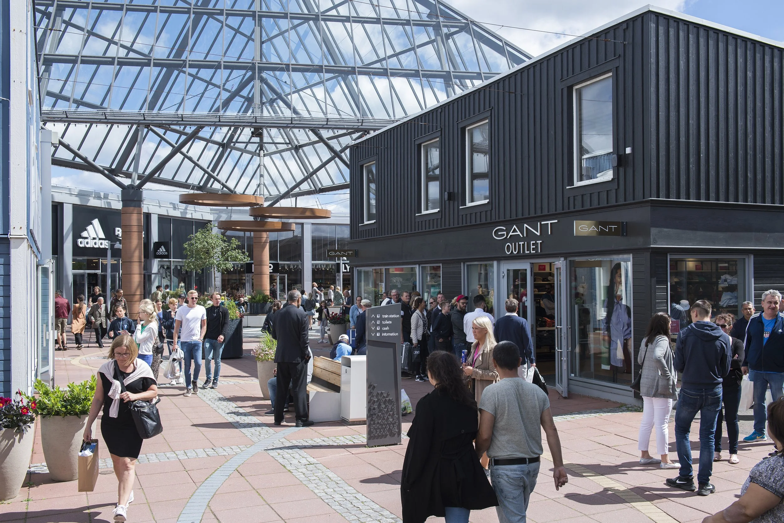 Crowd of people walking outside a shopping mall with a GANT Outlet store, a glass roof structure, and an information sign with icons for train station, toilets, cash, and information.