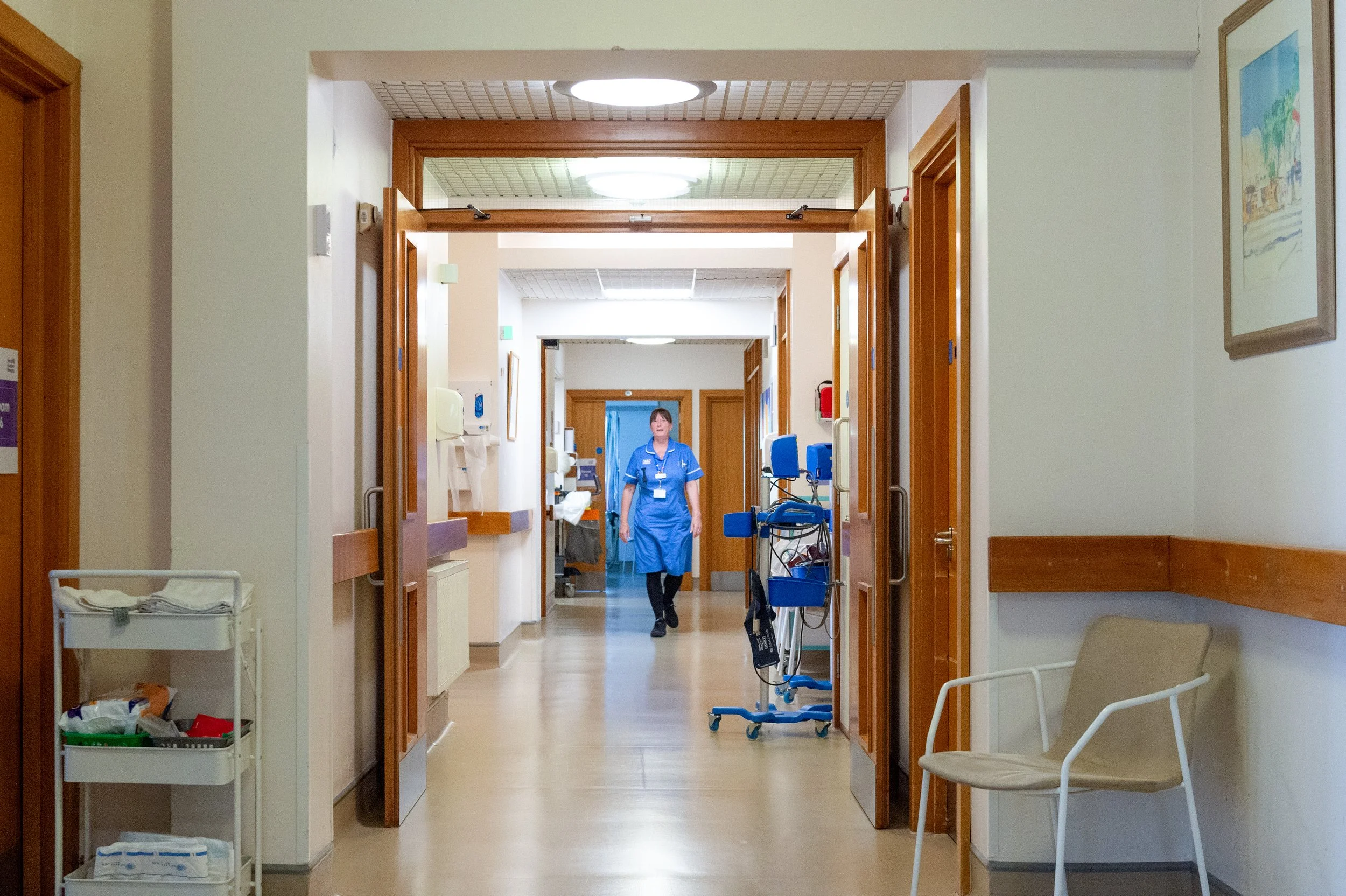 Hospital corridor with a nurse walking towards the camera, medical equipment, chairs, and framed artwork on the walls.