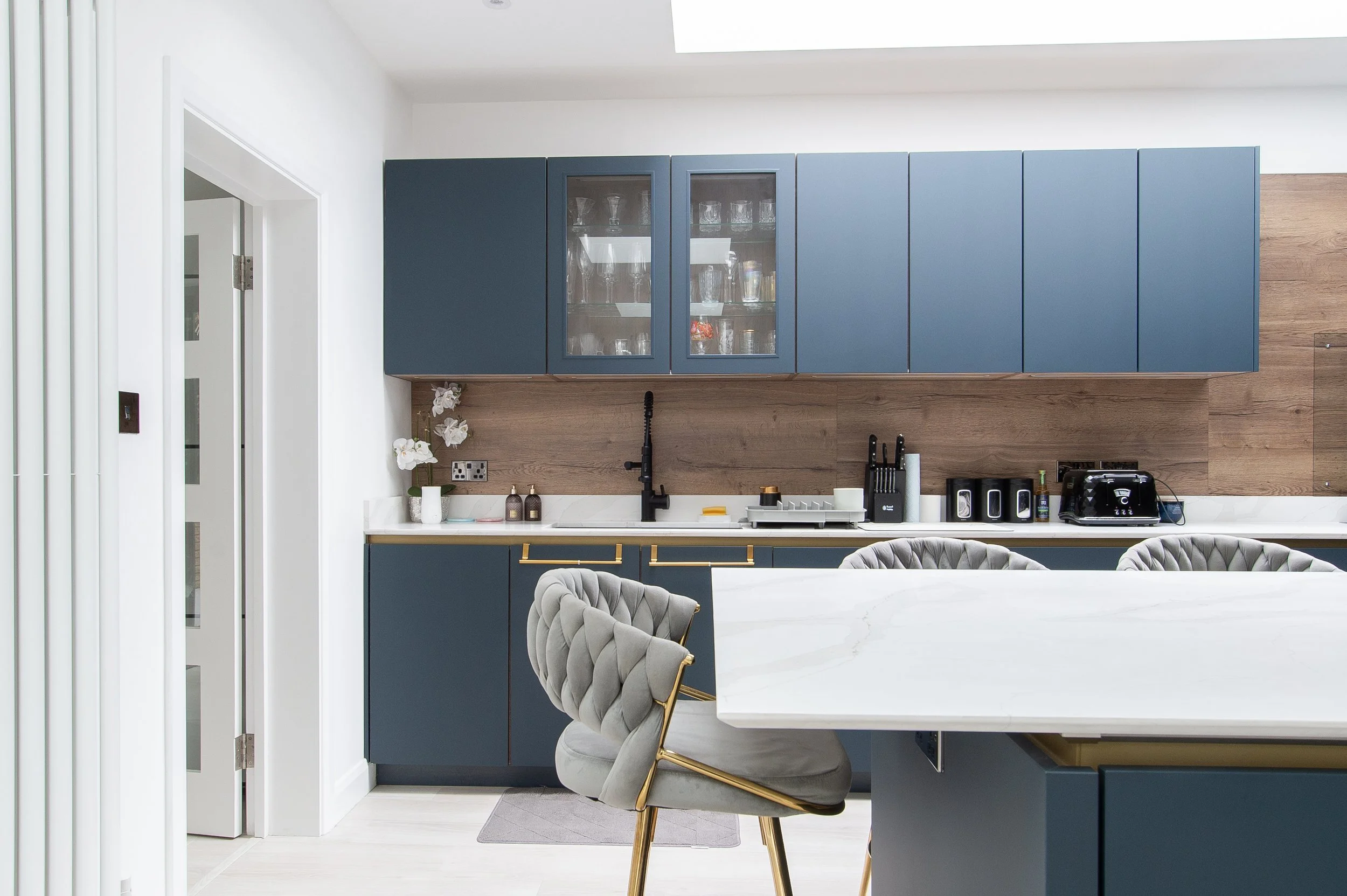 Modern kitchen with blue cabinets, glass-front upper cabinets, wooden backsplash, white countertops, black faucet, and various kitchen appliances on the counter. Part of a dining table and upholstered chair are visible in the foreground.