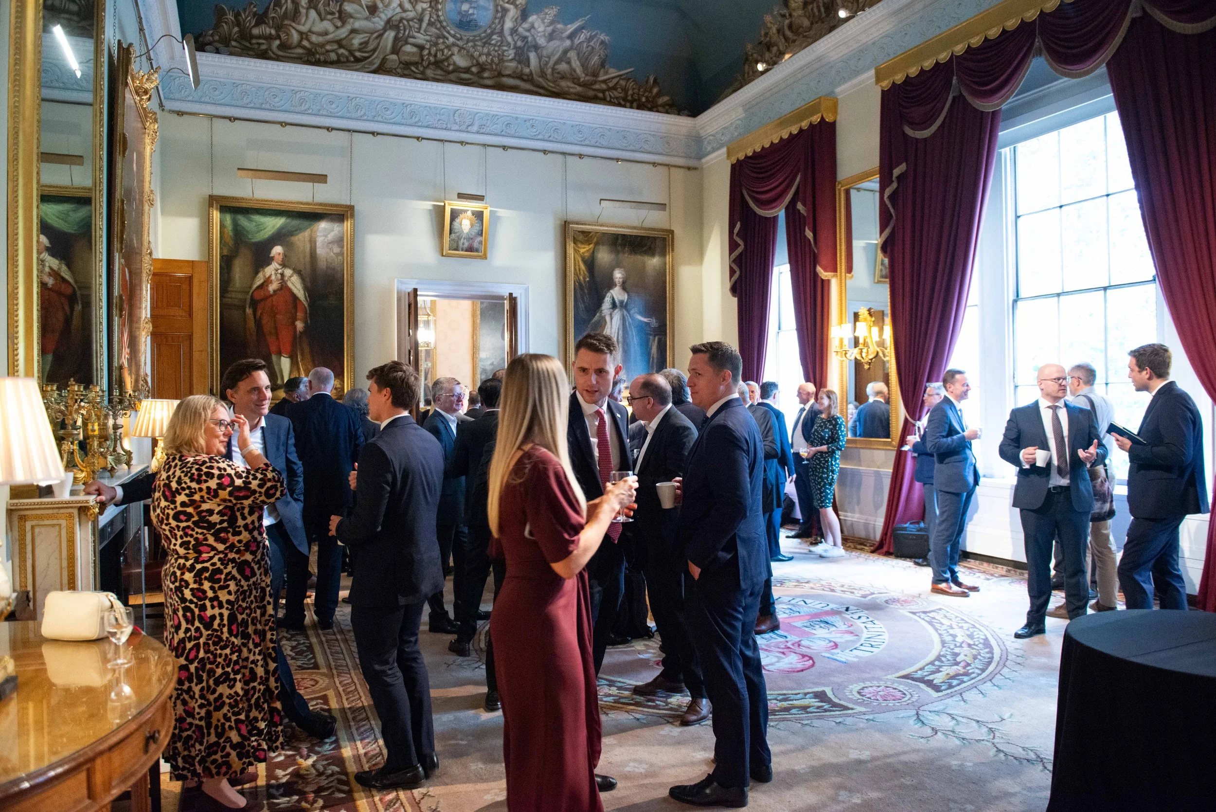 People socializing at a formal event in an ornately decorated room with large portraits, heavy drapes, and a detailed carpet.