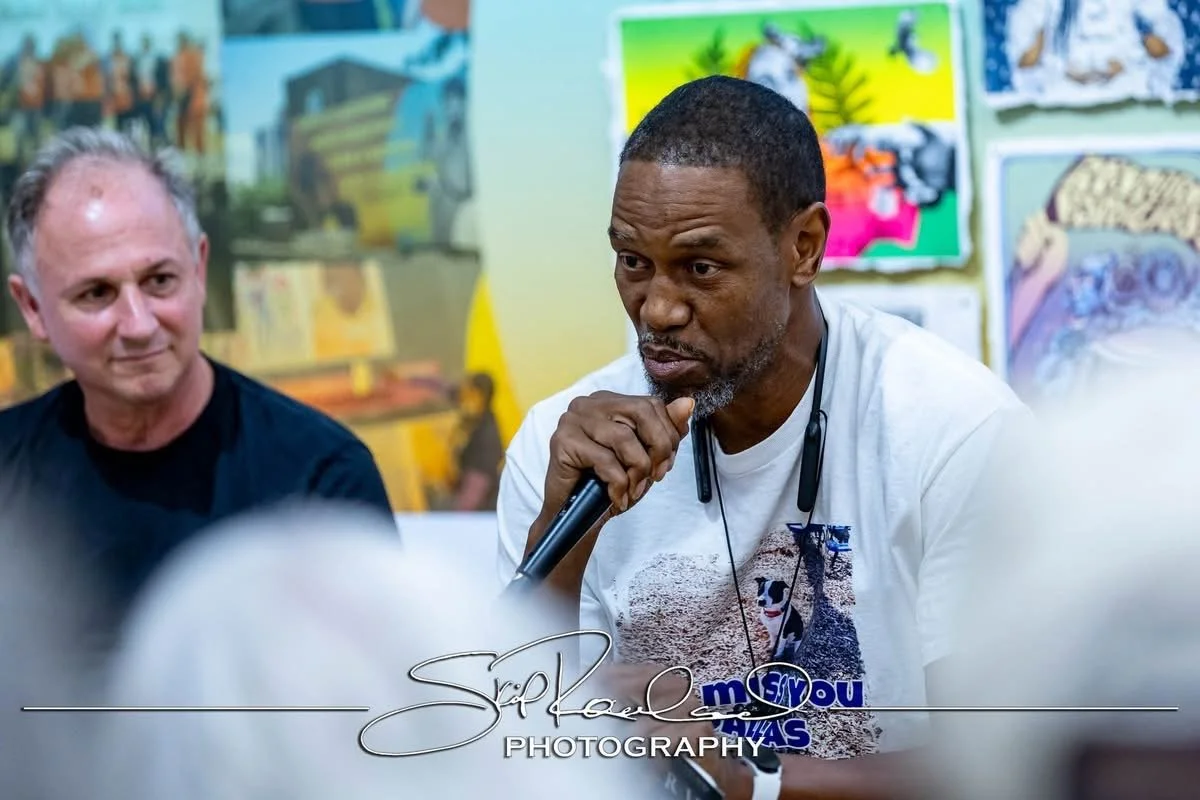 A man in a white T-shirt speaking into a microphone while others listen, with colorful artwork in the background.