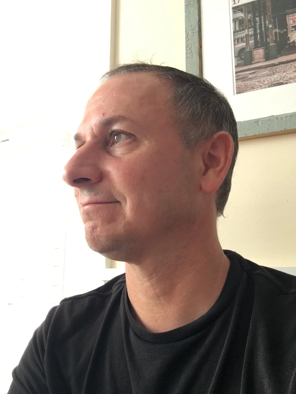 A middle-aged man with short dark hair and light skin looks to the left in a casual setting, with framed artwork or photographs on the wall behind him.