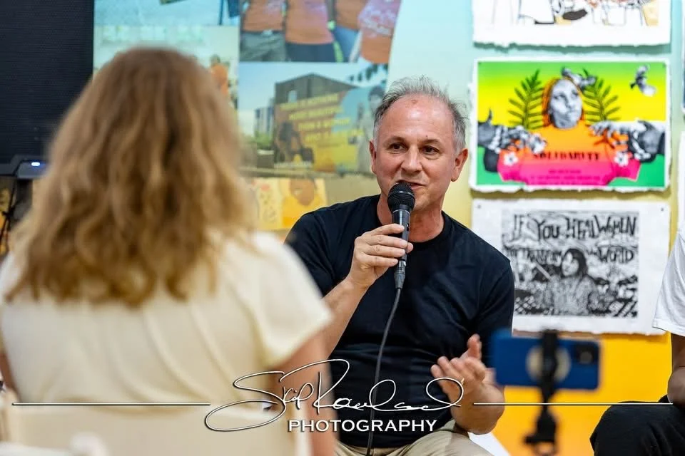 A man holding a microphone is speaking to a woman with red hair in a room decorated with colorful posters on the wall.