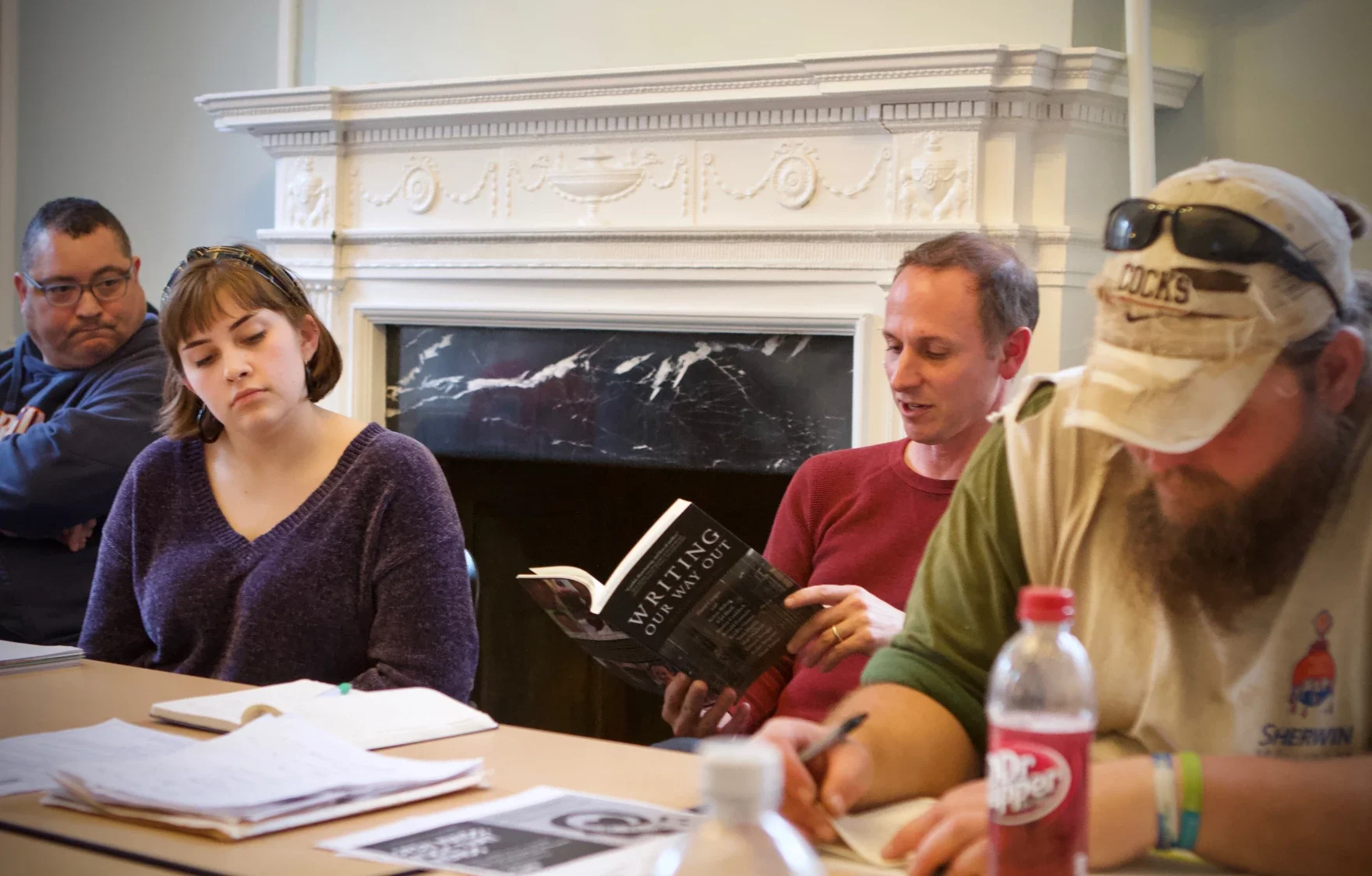 Group of four people sitting at a table, engaged in a discussion or meeting, with papers and drinks on the table, inside a room with a decorative fireplace.