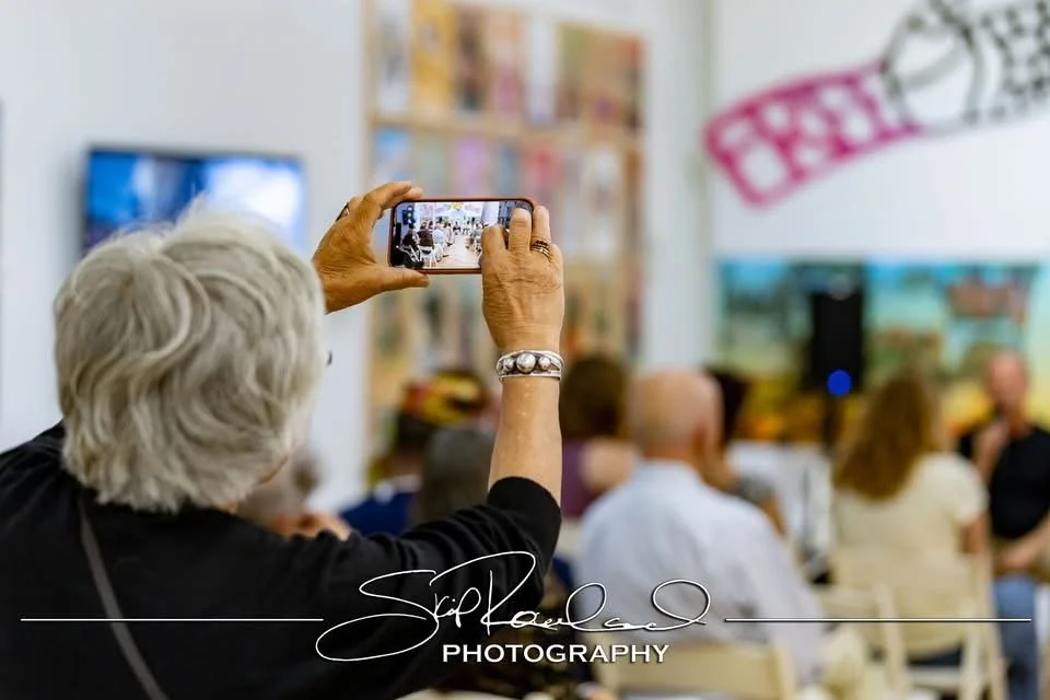 Older woman with white hair taking a photo with her smartphone at a gathering or event, with a blurred audience and presentation screen in the background.