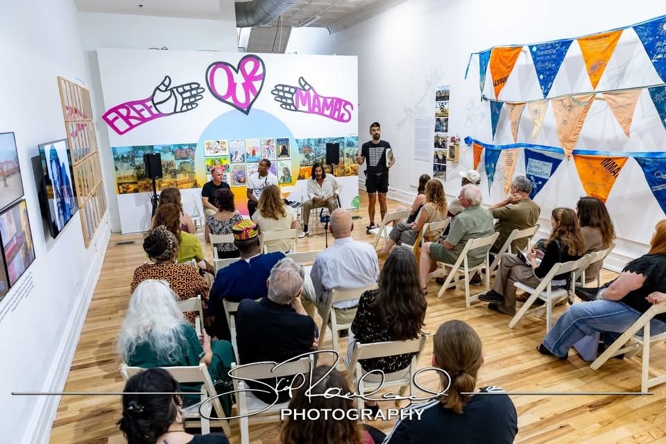 Community event with a panel of speakers sitting in front of an audience in a decorated room, featuring a large display with colorful signs that read 'CREC OF MAMAS' and a wall adorned with photos and orange and blue banners.