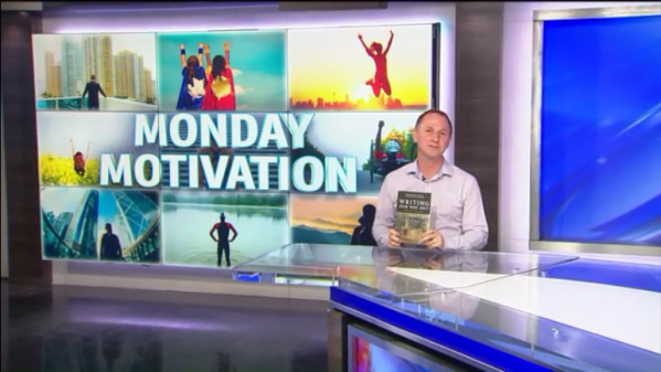 TV news studio with a man sitting at a news desk, holding a magazine, behind him a large screen displays 'Monday Motivation' with various images of people jumping and enjoying outdoor activities.