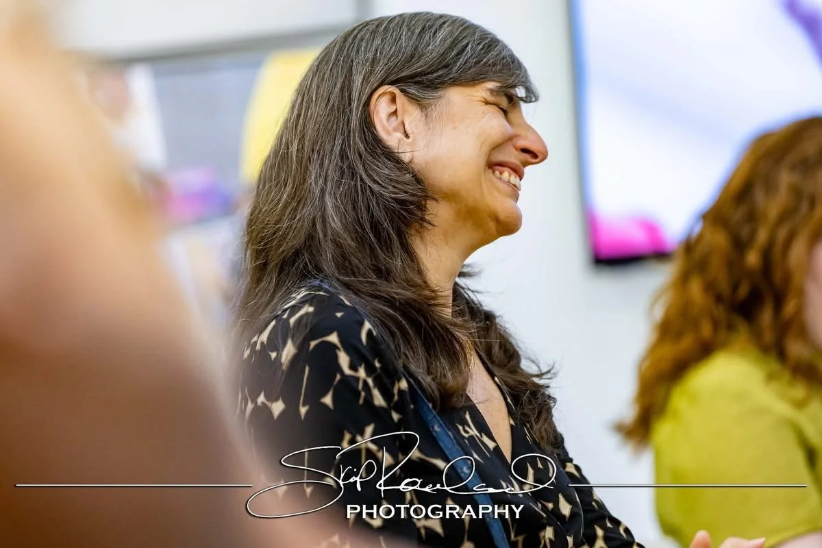A woman with long dark hair smiling and looking to the right during a meeting or presentation.