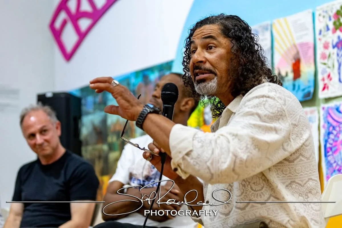 A man with curly hair and a beard speaking into a microphone during a panel discussion, with colorful artwork displayed in the background.