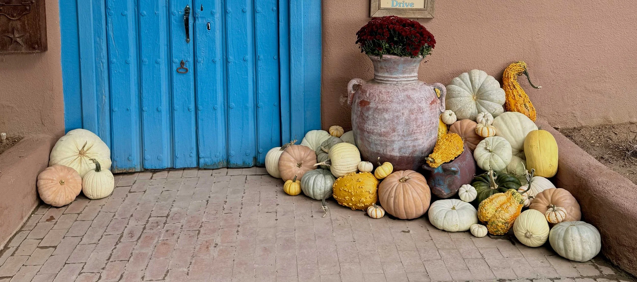 Display of pumpkins and gourds in various colors and sizes around a large, aged pink vase with red flowers on top, set against a blue wooden door and pink stucco wall.