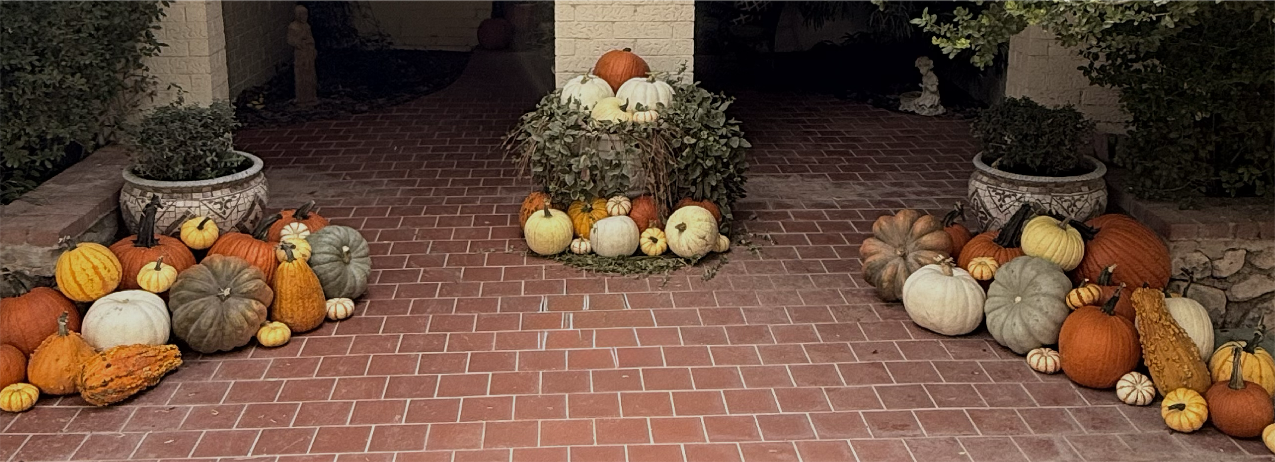 Arrangement of pumpkins and gourds of various sizes and colors, placed on a brick porch with potted plants and decorative statues, celebrating fall or Thanksgiving.