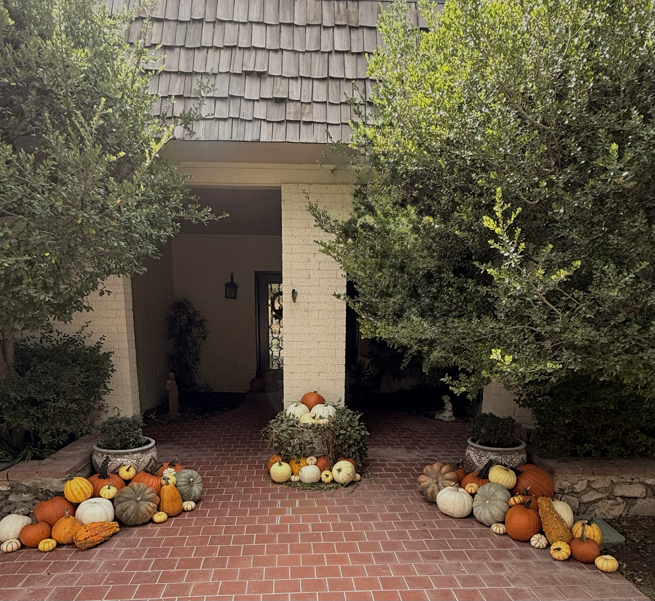 Front porch decorated with a large assortment of pumpkins and gourds, surrounded by trees and bushes, with a brick walkway leading to the entrance.