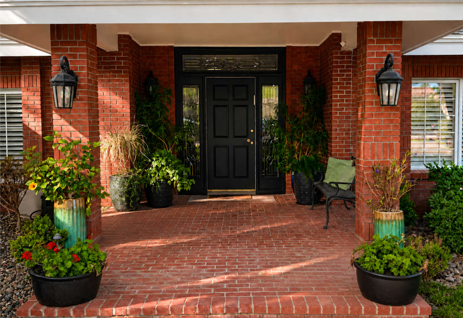 Front porch of a brick house with black door, surrounded by green plants in pots, a bench with a green cushion, and black wall-mounted lanterns.