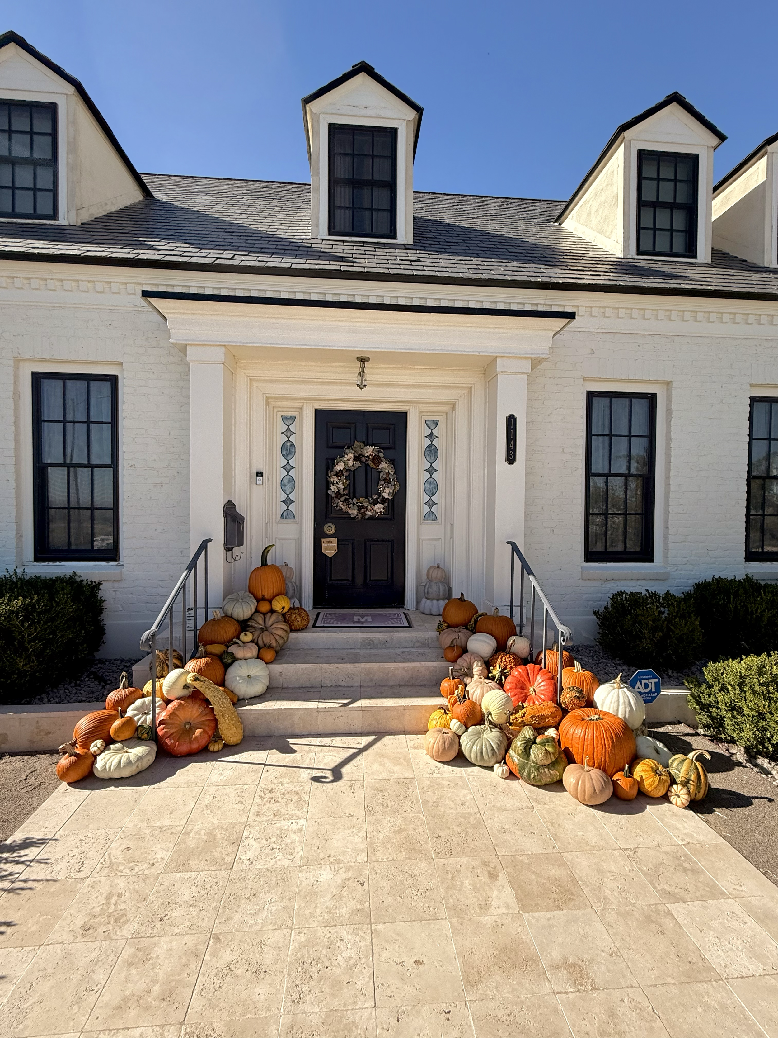 Front entrance of a white house decorated for fall with a door wreath and pumpkins arranged on the steps and sidewalk.