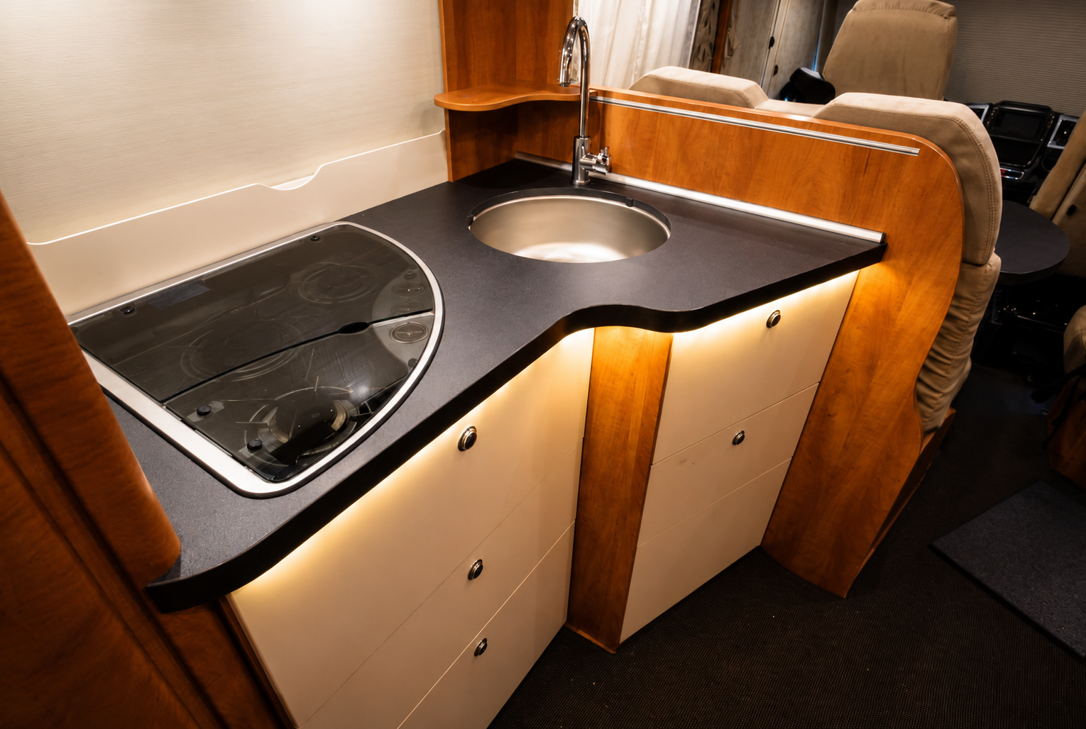 A compact kitchen area inside a recreational vehicle with a black countertop, a round sink with a faucet, a two-burner stove with a glass cover, wooden accents, and storage cabinets below.