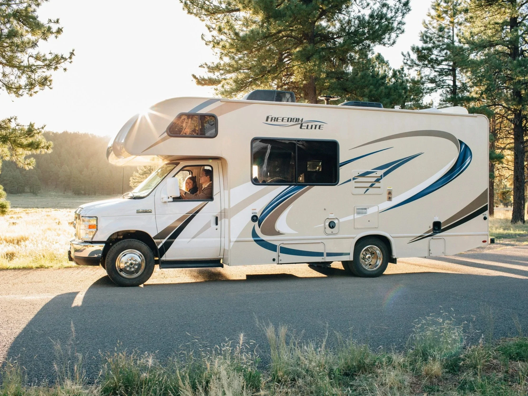 A white camper van parked outdoors in a wooded area during sunset, with two people inside smiling and looking out the window.