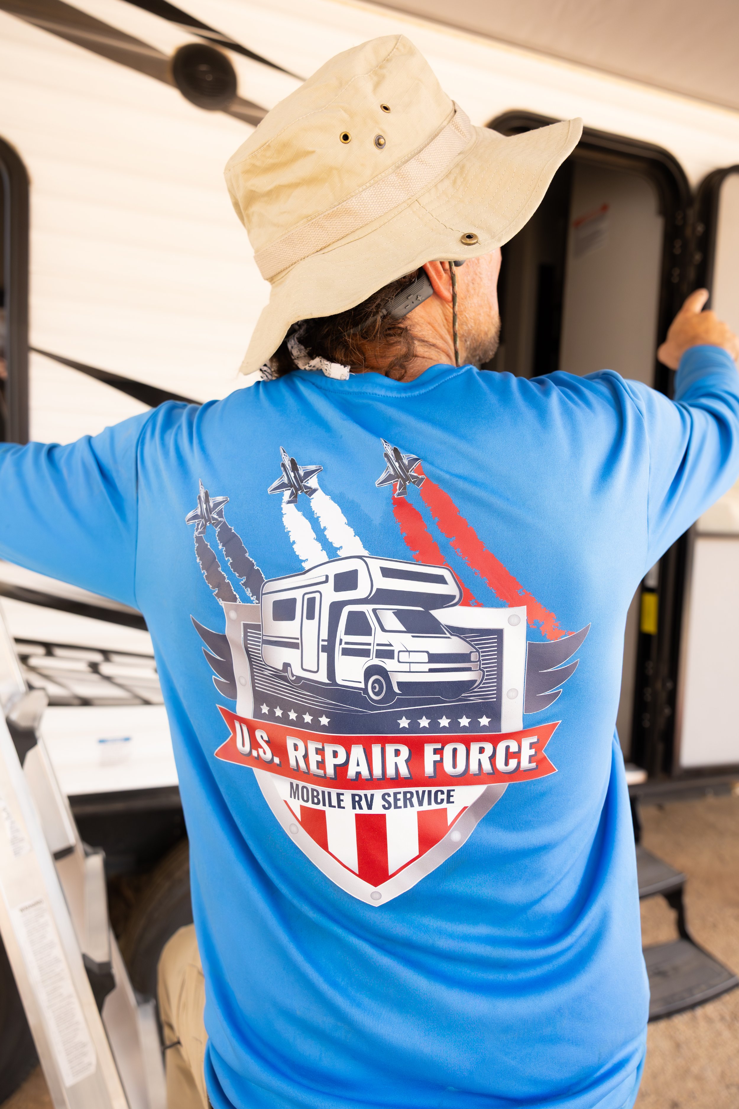 Person wearing a blue shirt with U.S. Repair Force logo, a large beige wide-brim hat, and listening to a device while working on a mobile RV service setup.
