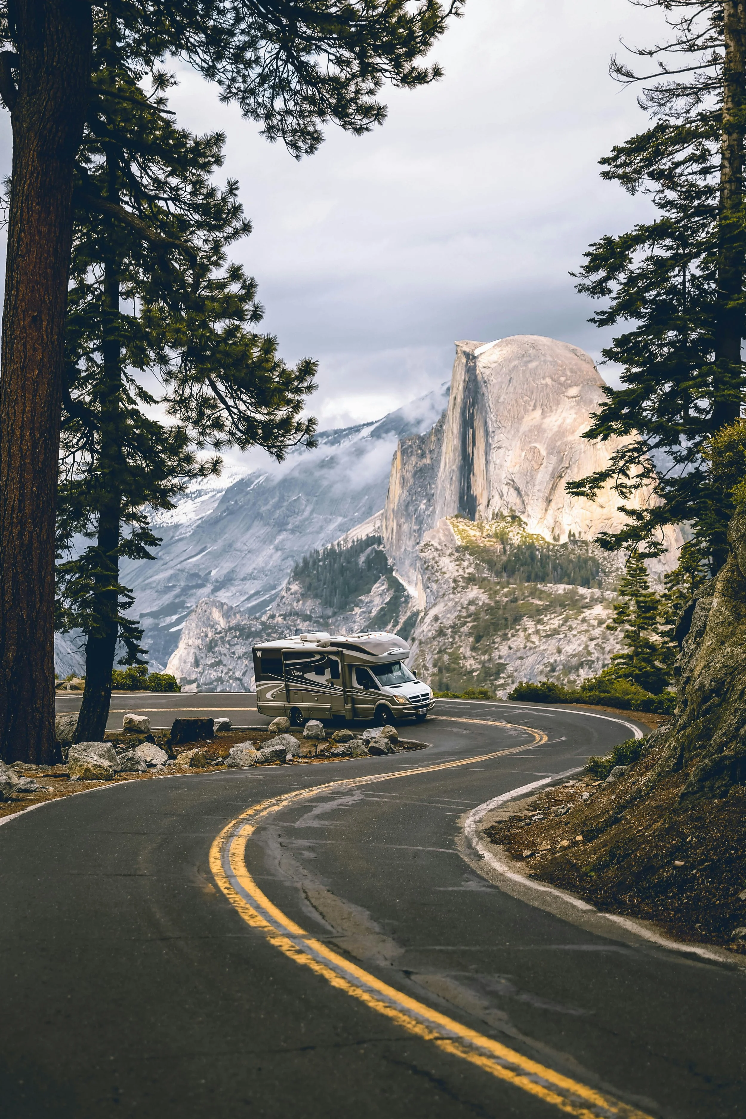 A gray camper van parked on a winding mountain road surrounded by tall pine trees, with large granite mountain and snow patches in the background.