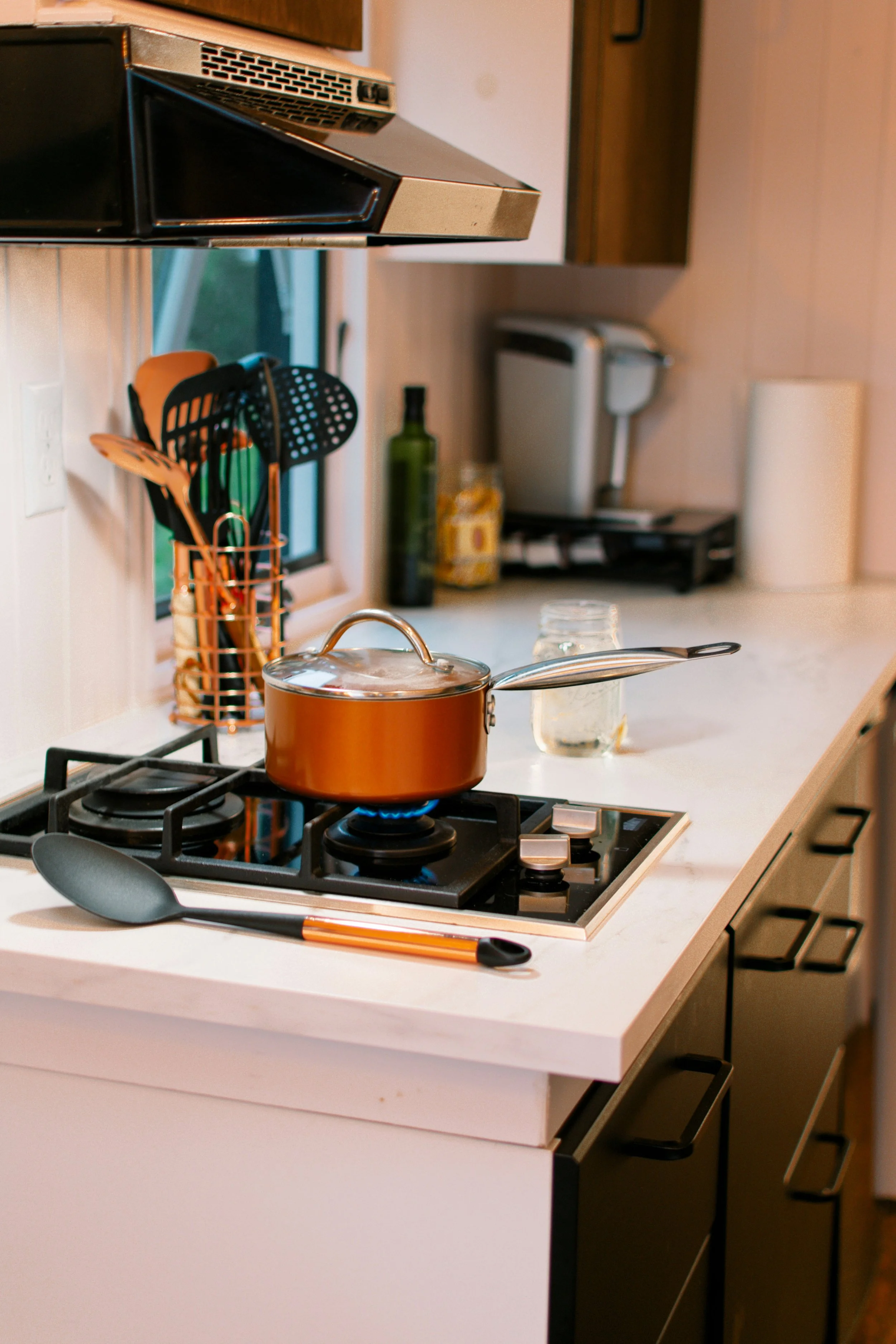 Close-up of a kitchen stove with a copper pot on burner, black spatula nearby, with kitchen utensils in a orange holder, in a cozy kitchen with cream counter and appliances.
