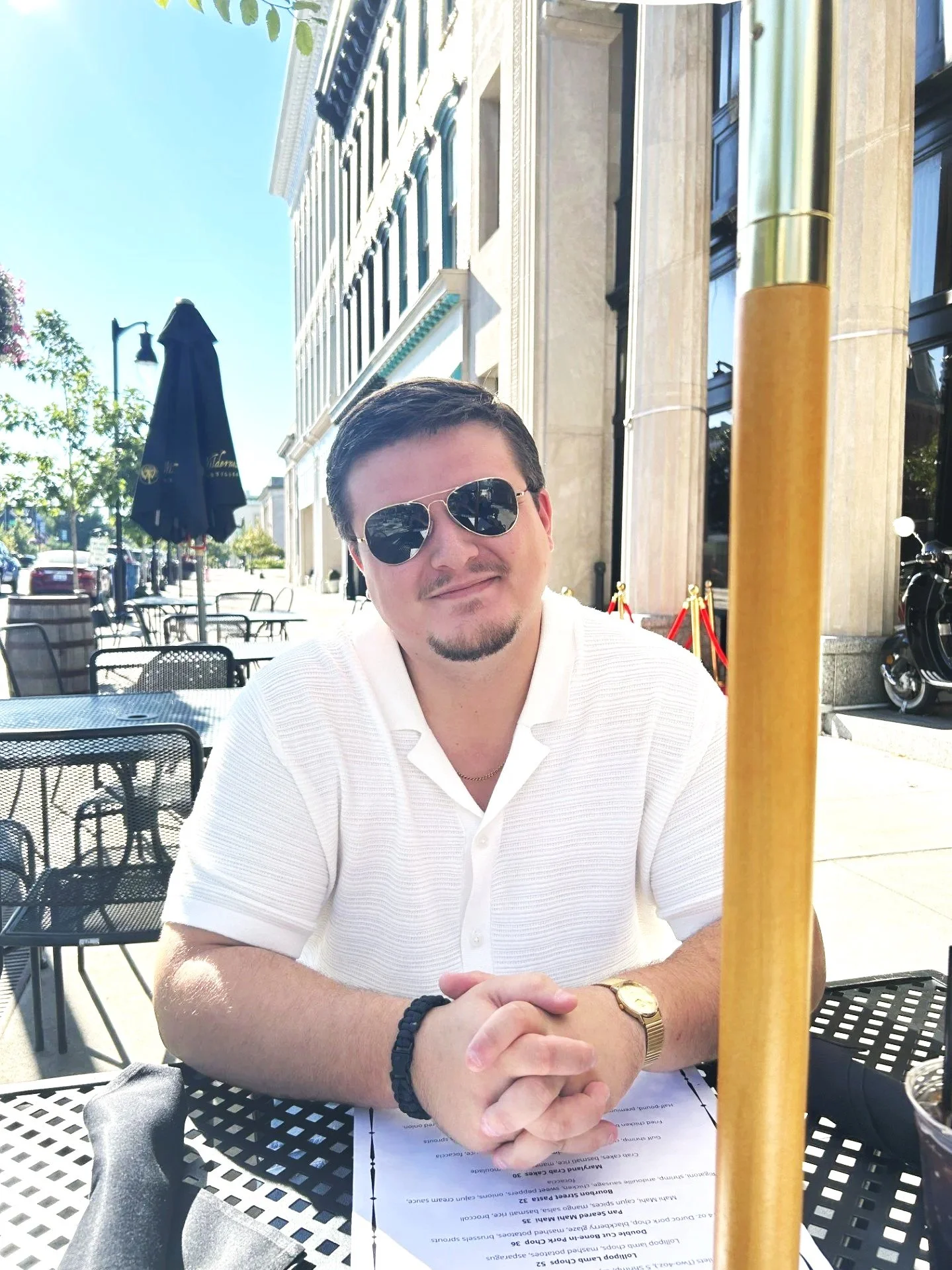 A man sitting at an outdoor cafe, wearing sunglasses and a white collared shirt, with a street scene and building in the background.
