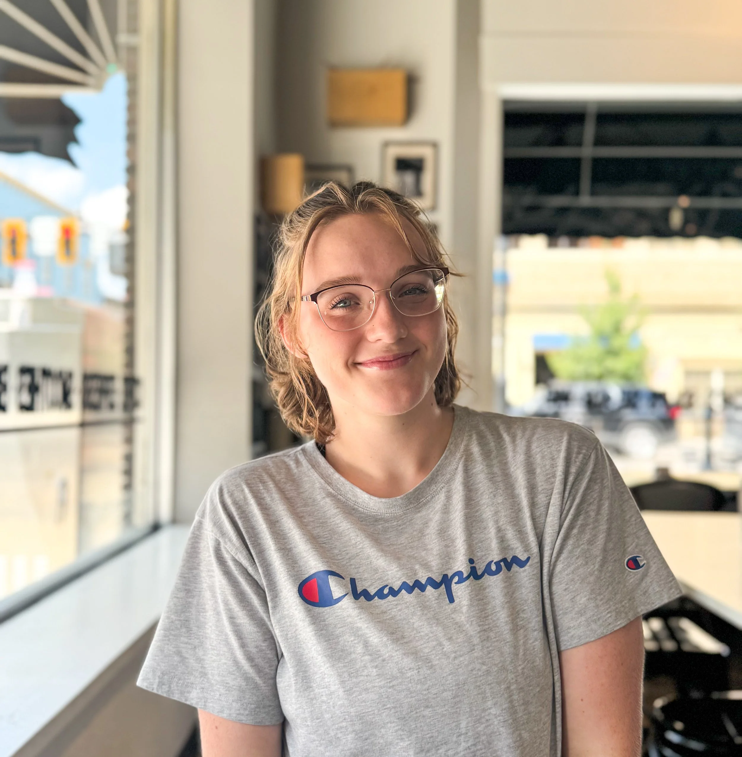 Close-up of a young woman with glasses, smiling, wearing a gray t-shirt with the Champion logo, inside a brightly lit store or shopping mall with large windows and a display outside.