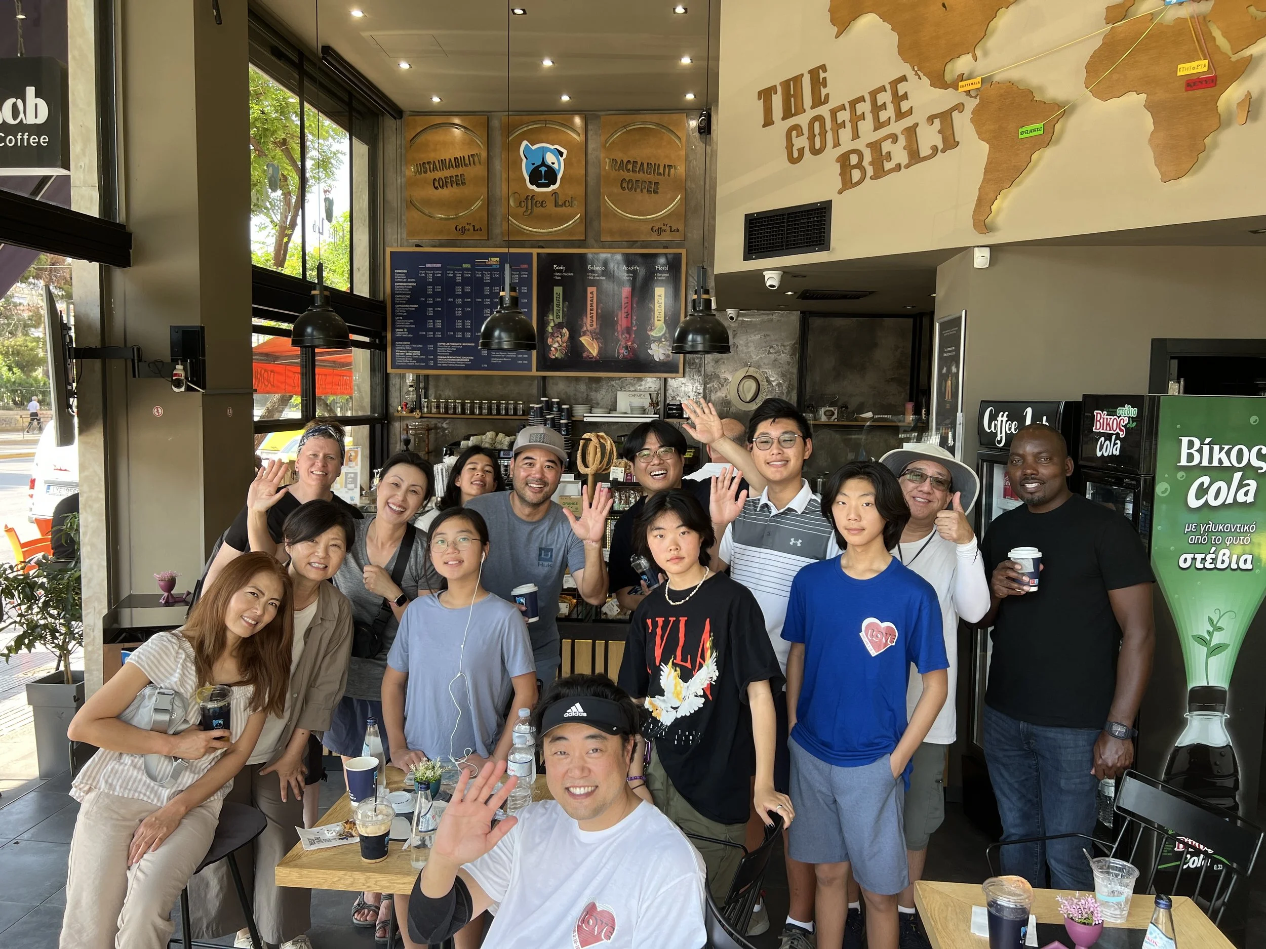 Adults and teenagers from the Saddleback Church VBS team smile and wave for a group photograph inside a café in Athens.